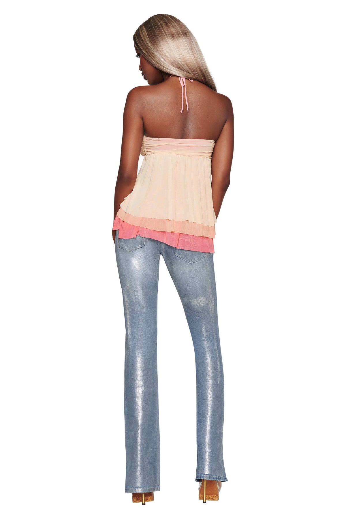 A woman with long, straight blonde hair stands with her back to the camera against a white background. She is wearing the I.AM.GIA IDALIA TOP in Raspberry Multi—a light pink mesh halter neck top embellished with multicolour ruffles—paired with shiny, light blue flared jeans and high heels.
