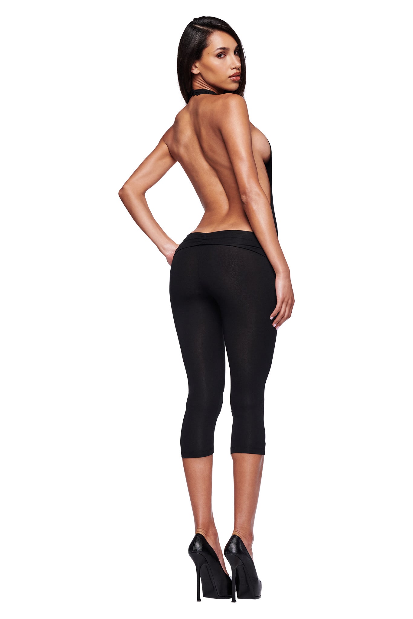 A woman faces away, glancing over her shoulder in the I.AM.GIA KAIRA JUMPSUIT - BLACK. The backless capri jumpsuit features a ruched waistband and is paired with high heels against a plain white background.