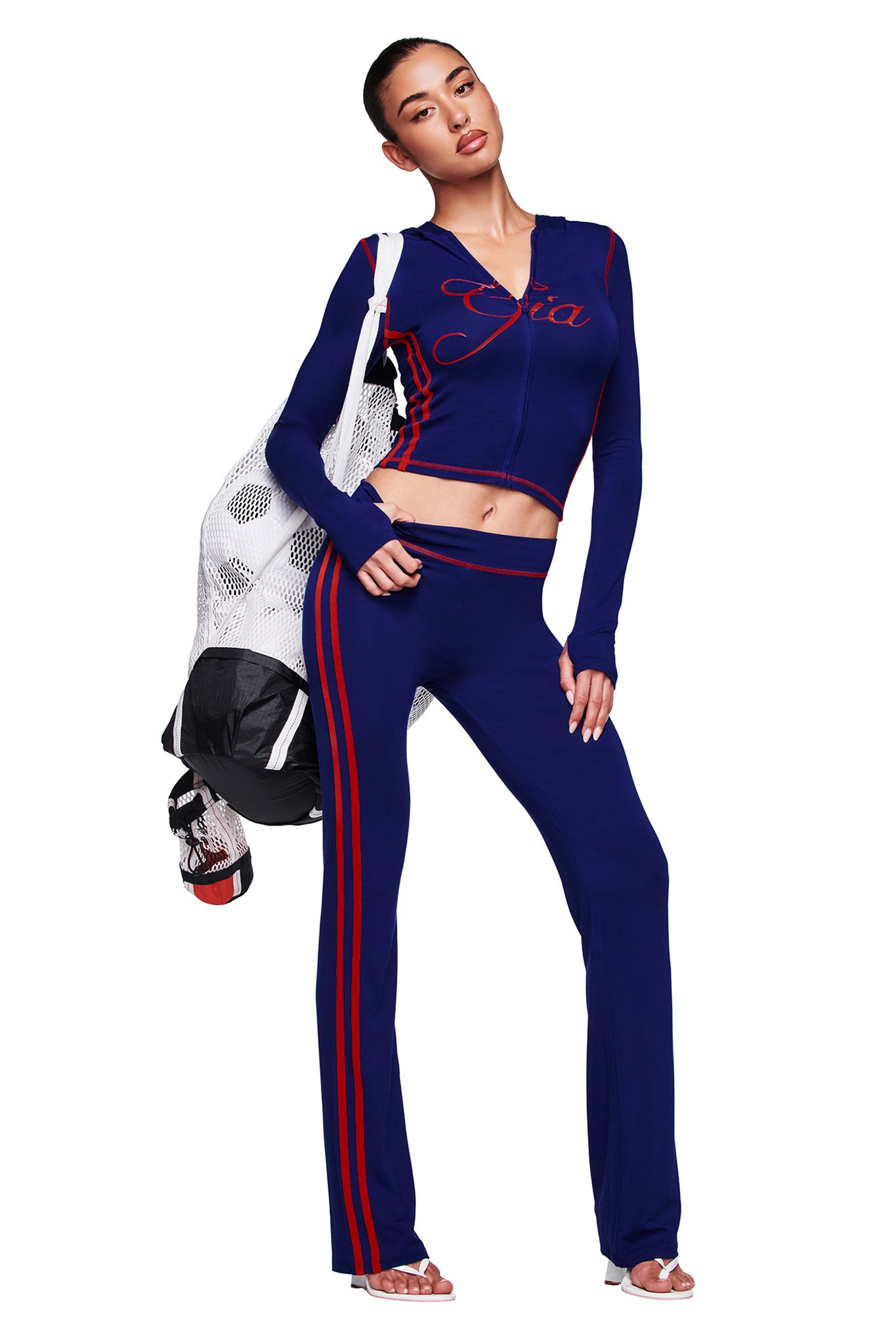 A woman stands confidently in I.AM.GIA's BLARE SPORT TRACKPANT - NAVY, featuring red stripes. She pairs it with a blue crop jacket, white mesh bag, and white sandals, posing against a white background.