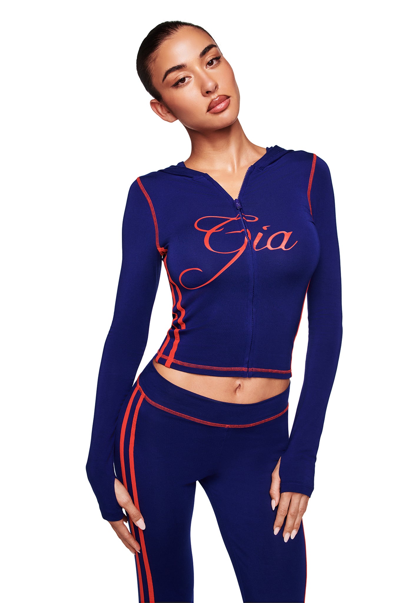 A woman models the I.AM.GIA BLARE TWO-PIECE SPORT HOODIE in navy, featuring red accents and "Gia" on the chest, paired with matching navy pants with red side stripes, all made from buttery-soft fabric, against a white background.
