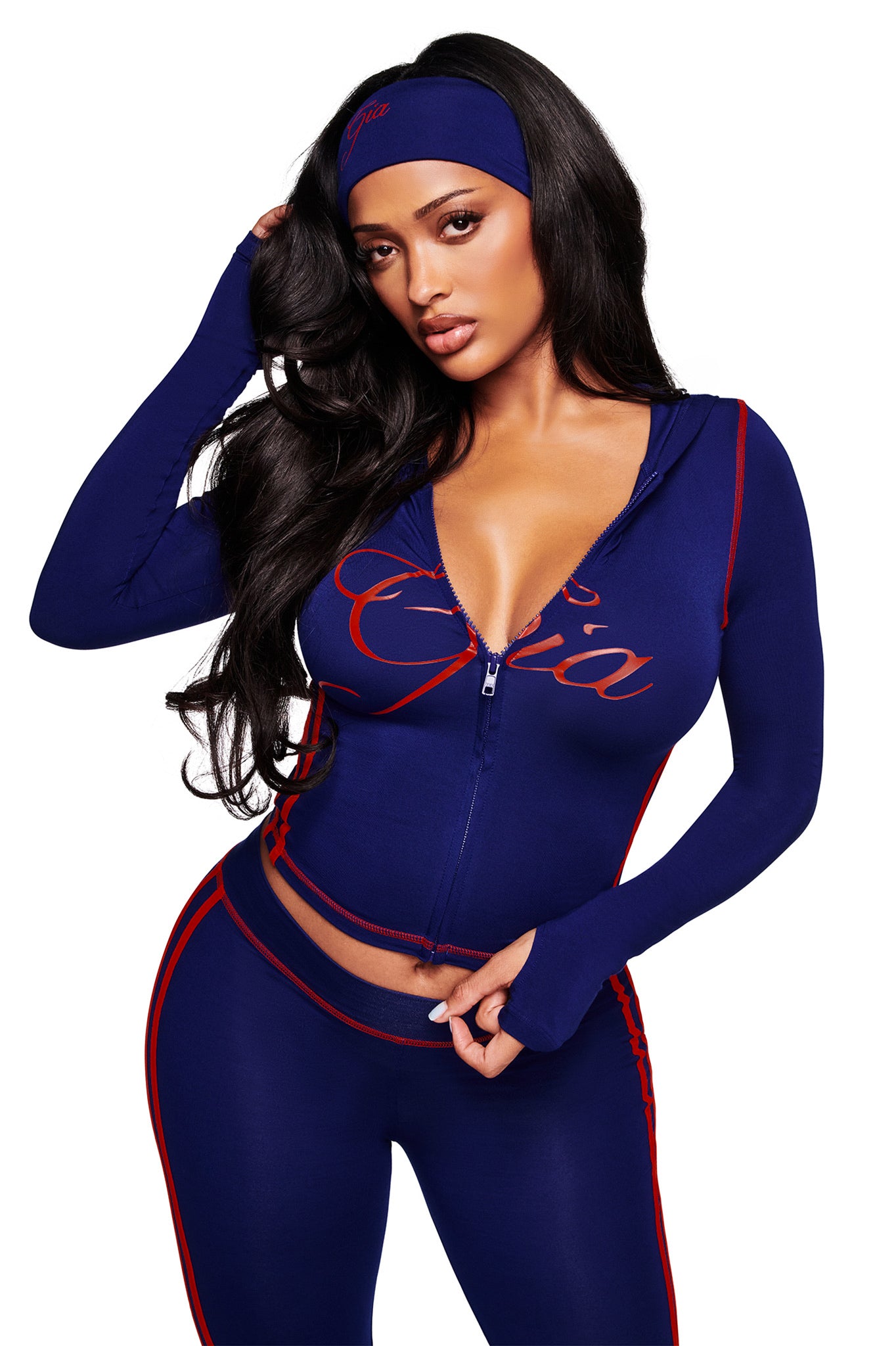 A woman with long dark hair poses in the I.AM.GIA BLARE SPORT HOODIE AND HEADBAND in navy, the buttery-soft hoodie partially unzipped as she touches her head and rests her other hand on her waist.