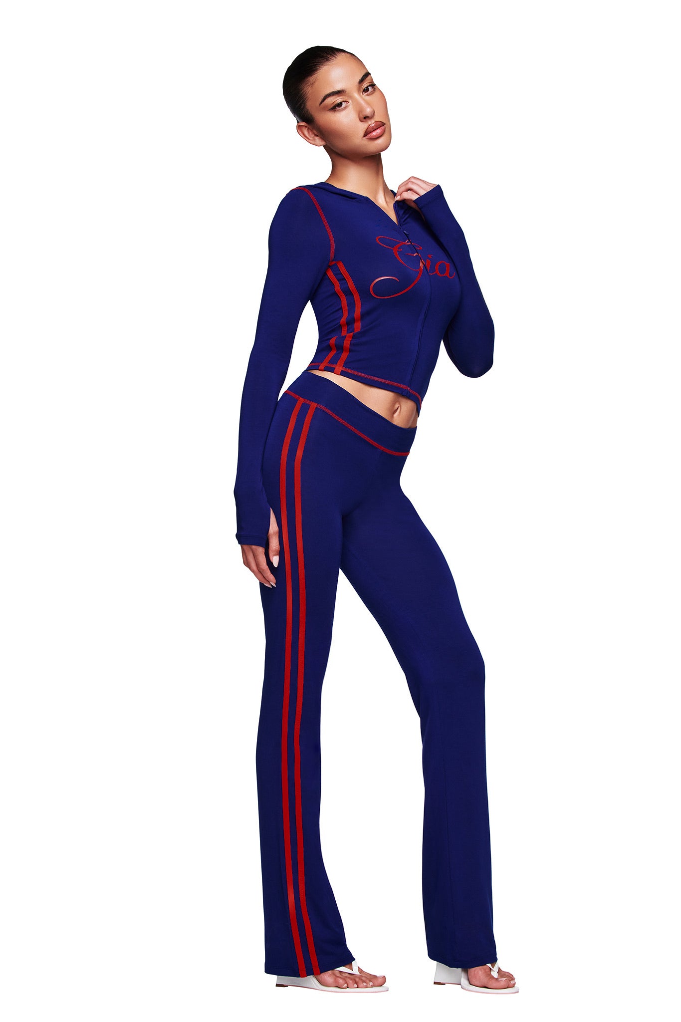 A woman models the I.AM.GIA BLARE SPORT TRACKPANT in navy—a fitted two-piece set with red stripes, script on the top, long sleeves, and flared pants—paired with open-toe heels against a white background.
