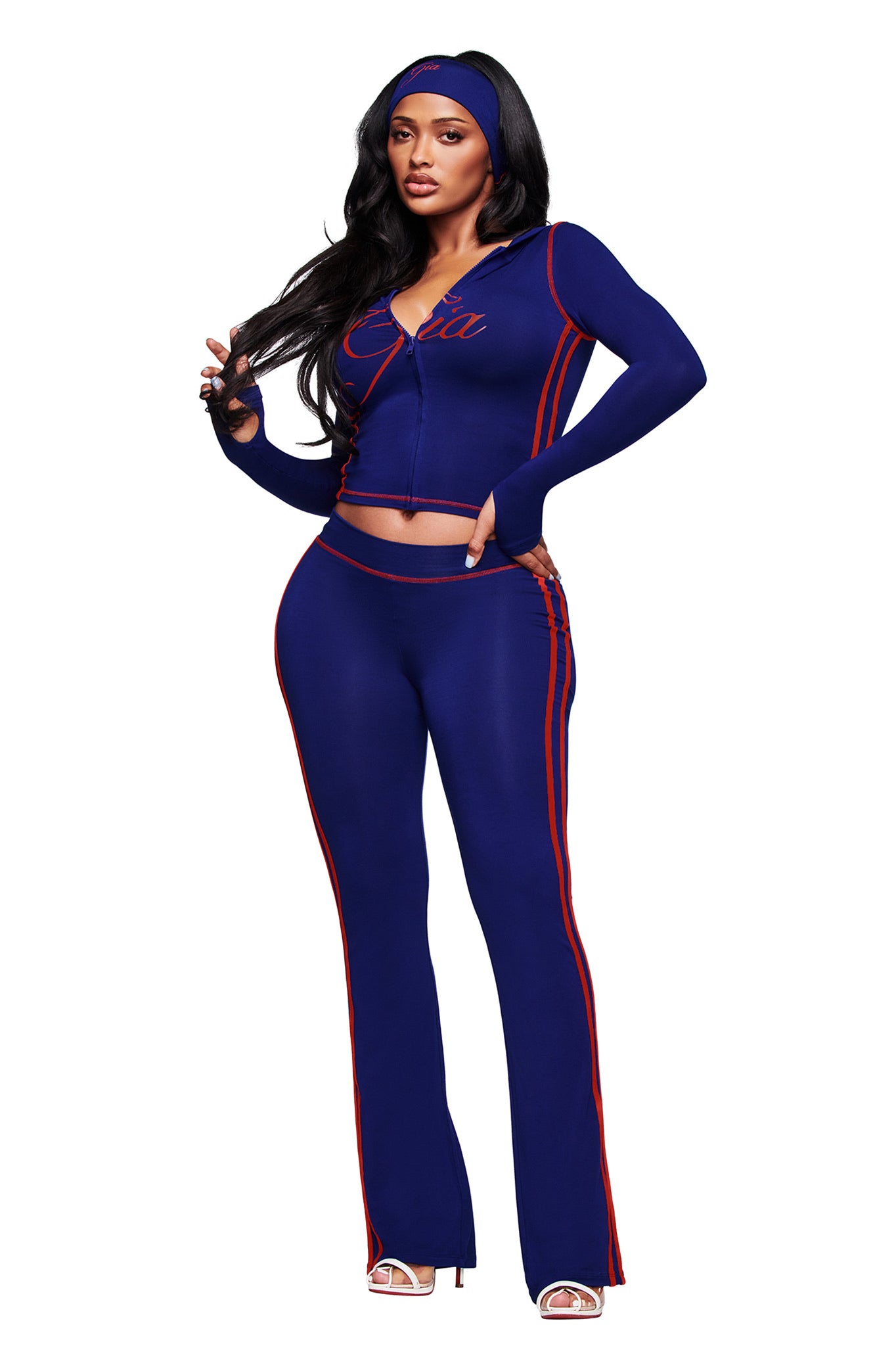 A woman poses in the I.AM.GIA BLARE SPORT TRACKPANT in navy, styled with a sporty blue zip-up jacket with red stripes, open-toe heels, a matching headband, and long dark hair. She stands with one hand touching her hair.