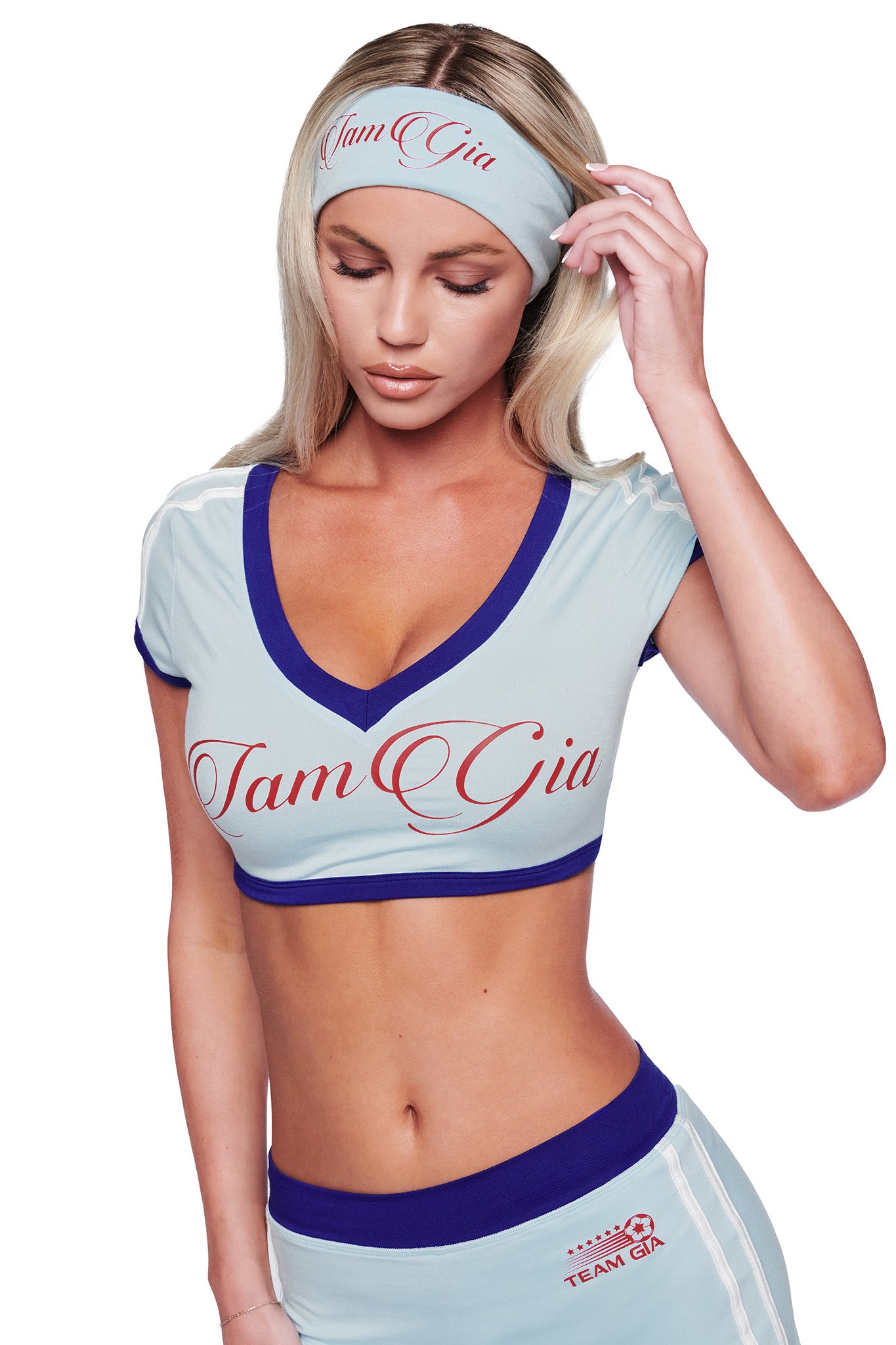 A woman in a baby blue BLARE SPORT HEADBAND by I.AM.GIA, along with a matching crop top and athletic shorts, adjusts her headband while looking down. The background is white.