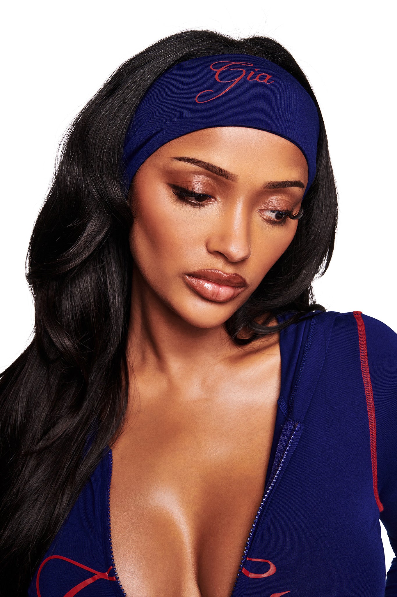 A woman with long dark hair wears the I.AM.GIA BLARE SPORT HEADBAND in navy, embroidered with "Gia" in red script, and a matching blue top with a partially unzipped front. She looks down calmly against a white background.
