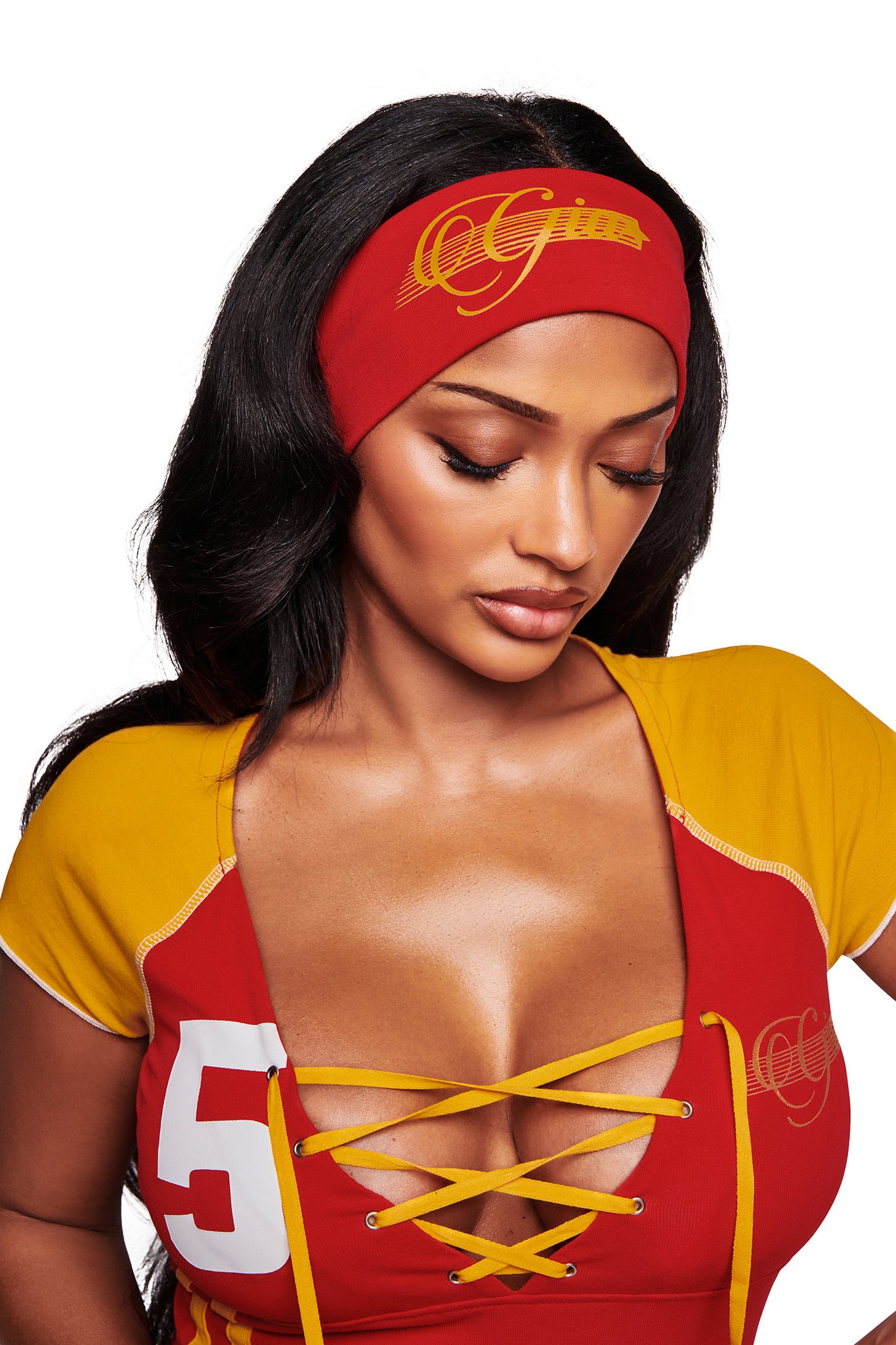 A woman with long black hair wears a red and yellow lace-up top featuring the number 5 and "OG," paired with the I.AM.GIA NOVIA SPORT HEADBAND - RED that has "OG Girls" written on it, as she looks down.