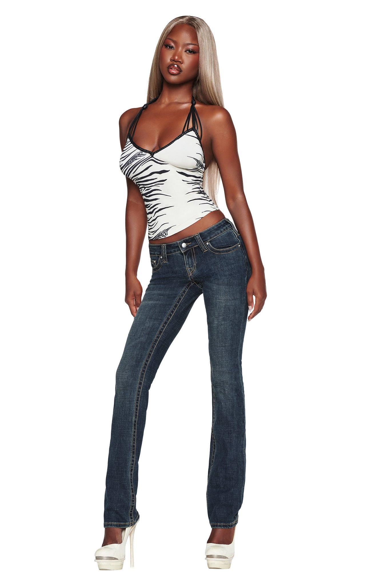 A woman with long blonde hair wears a white and black halter top, the I.AM.GIA BIANKA JEAN in blue, and white high heels, posing confidently against a white background.