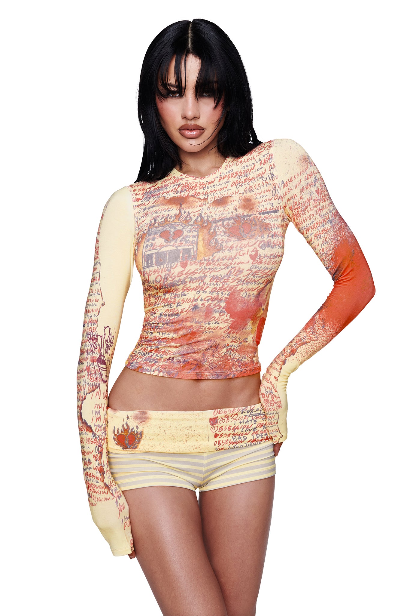 A woman poses with one hand on her hip against a white background, wearing the I.AM.GIA T-REX BLARE 2.0 LONG SLEEVE TEE in yellow—a fitted, long sleeve crewneck top featuring handwritten text and abstract red-orange patterns—paired with matching shorts.