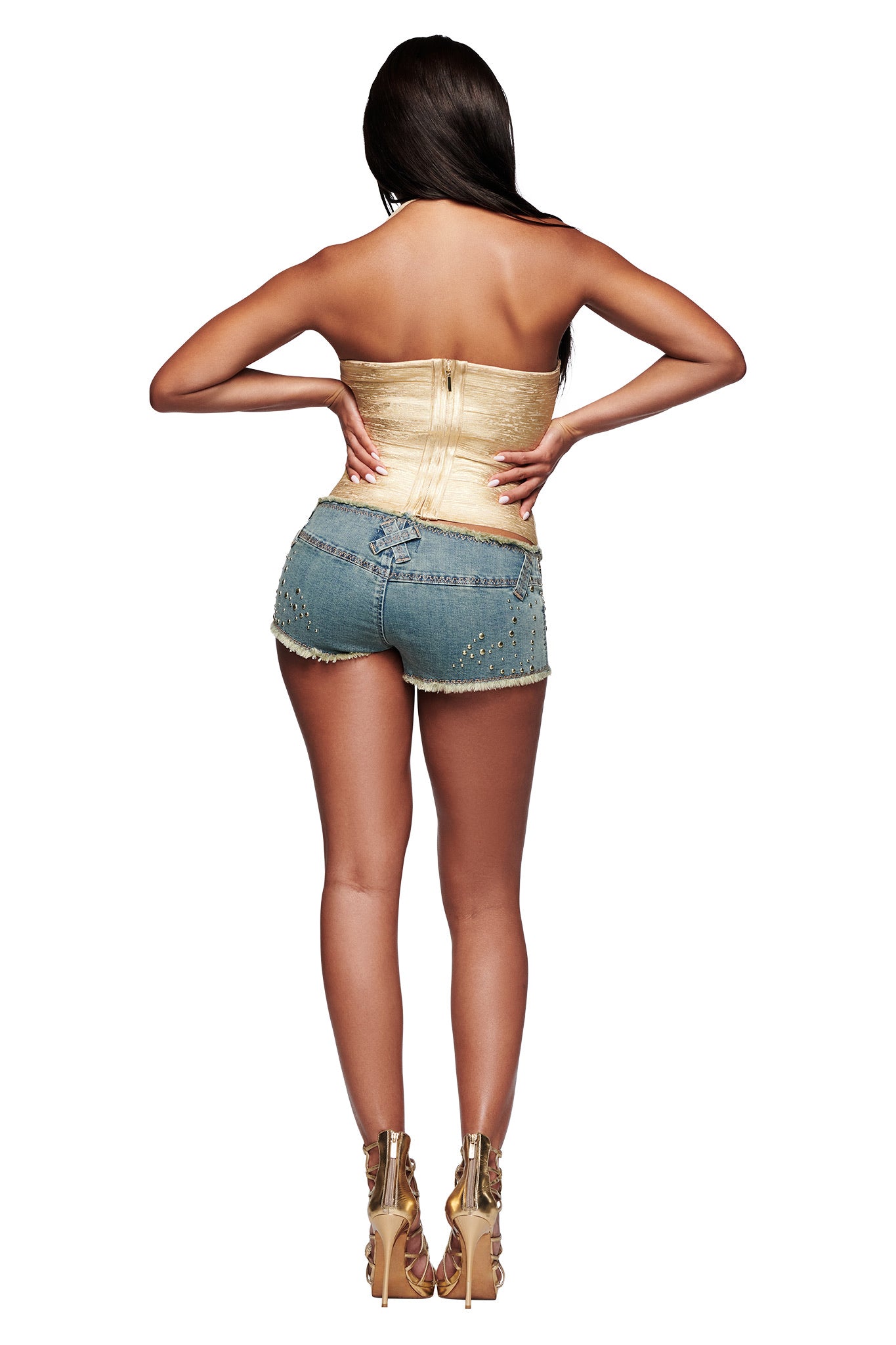 A woman with long dark hair stands facing away in a white setting, wearing the I.AM.GIA AIDEN PLATINUM BANDAGE TOP in gold paired with studded denim shorts and high heels, hands resting on her lower back.