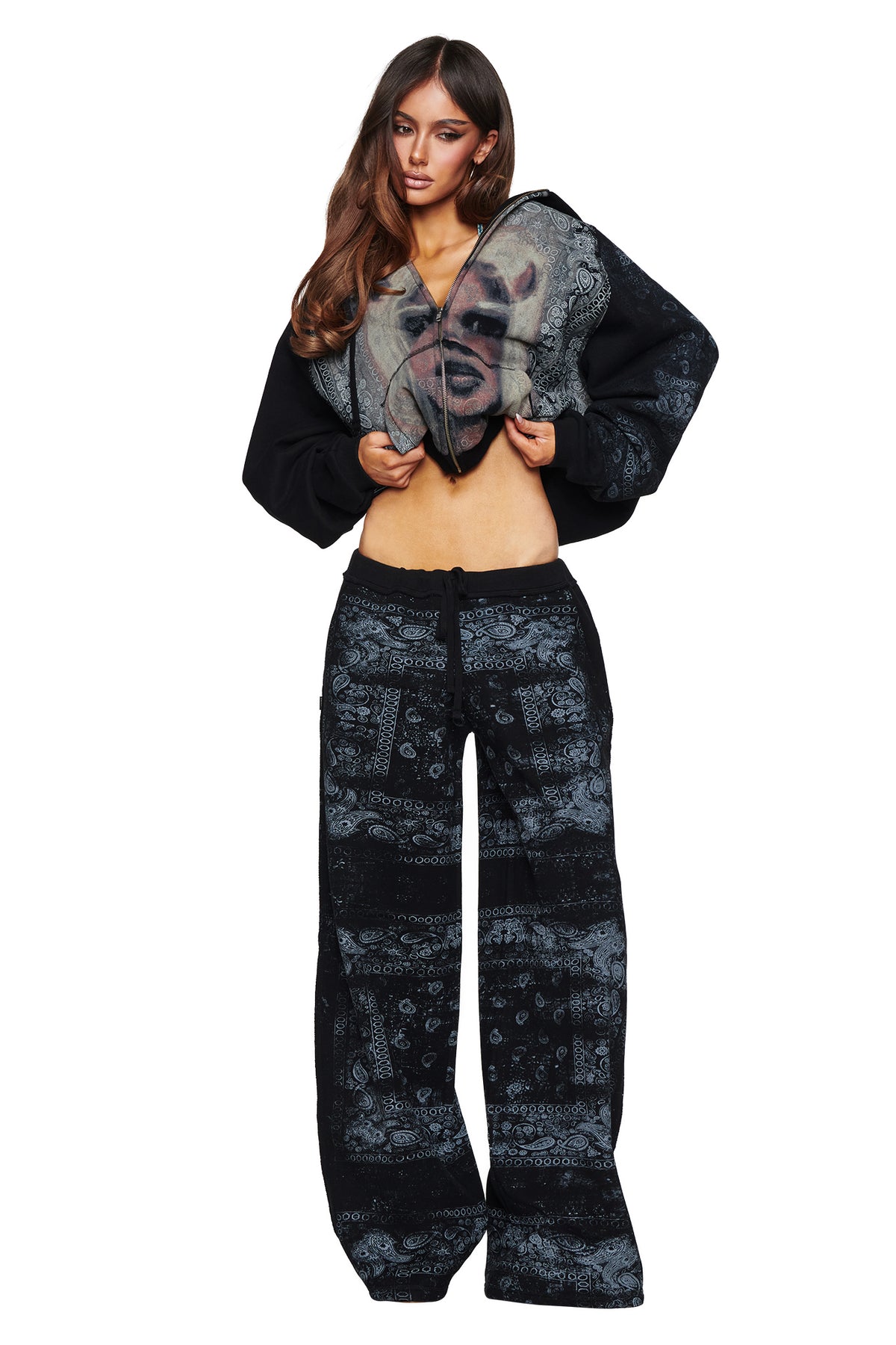 A woman stands against a white background in the I.AM.GIA T-REX TRACKPANT - BLACK BANDANA, pairing the bold black patterned pants with a cropped hoodie that features an oversized face graphic; as she lifts the hoodie to reveal her midriff, she makes a striking contemporary street style statement.