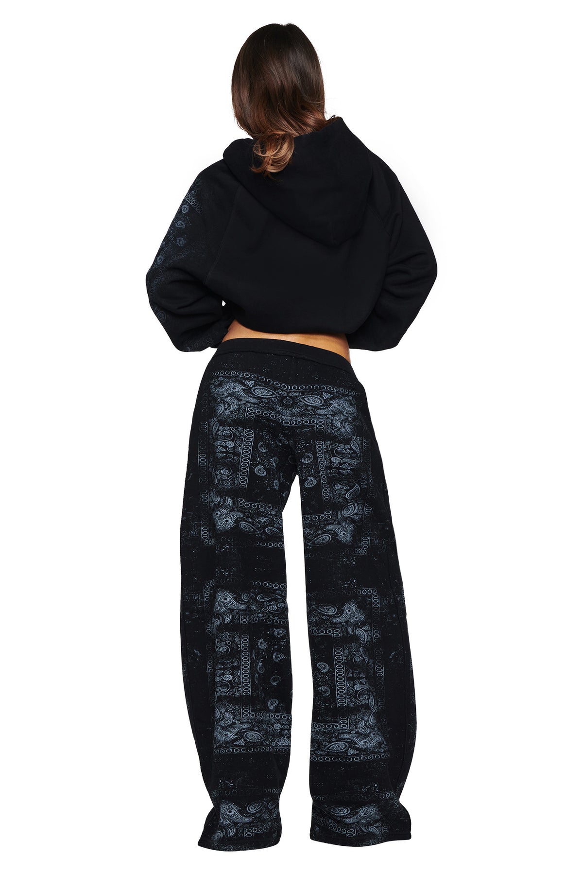 A person stands facing away, wearing the I.AM.GIA T-REX ZIP THRU HOODIE in Black Bandana paired with loose black pants featuring a gray paisley pattern, channeling effortless street style against a white background.