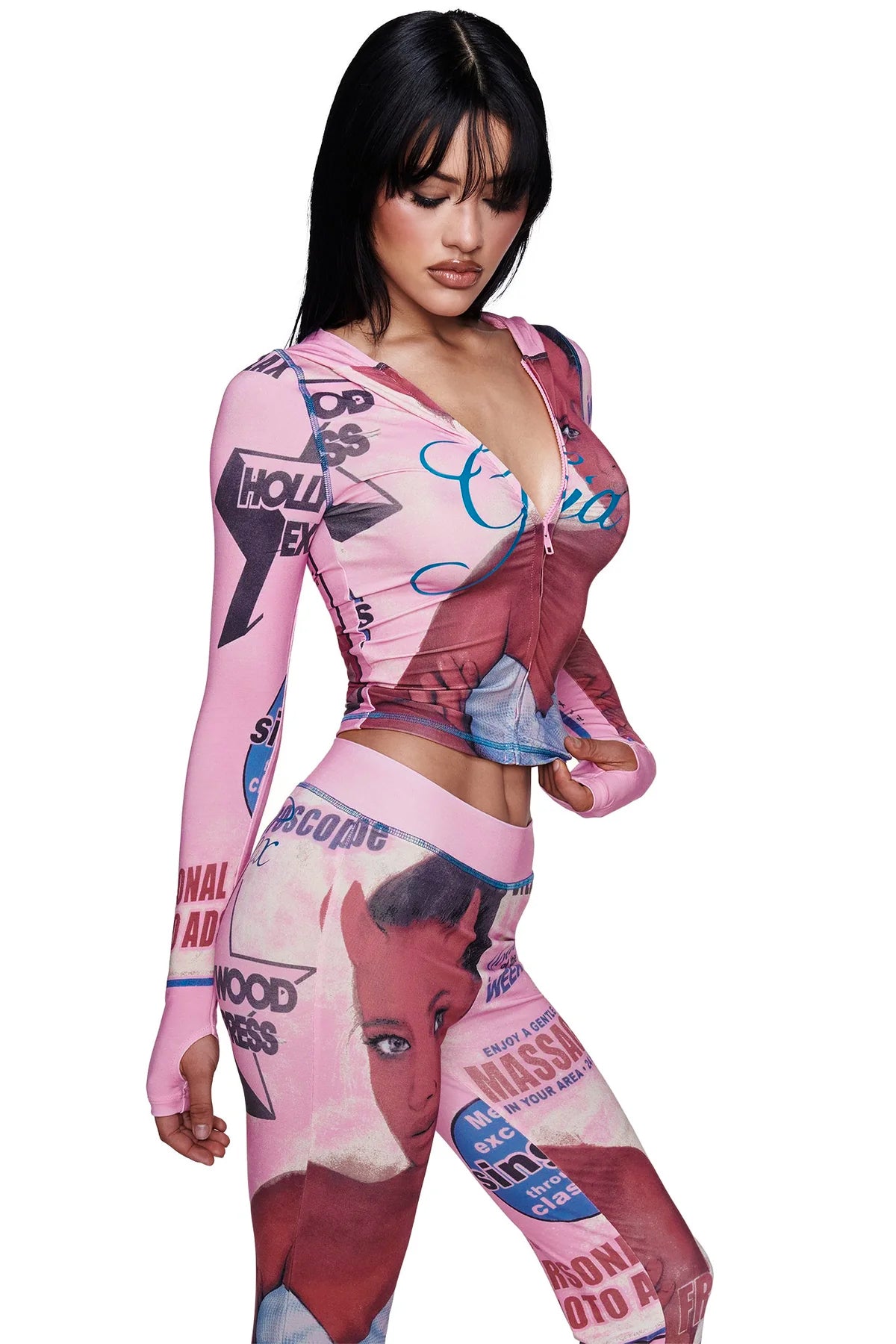 A woman with long dark hair wears the I.AM.GIA T-REX BLARE TRACKPANT in pink, styled as part of a fitted and colorful outfit featuring retro-inspired graphic prints, faces, and bold text. She stands against a plain white background with her hand on her hip, gazing downward.