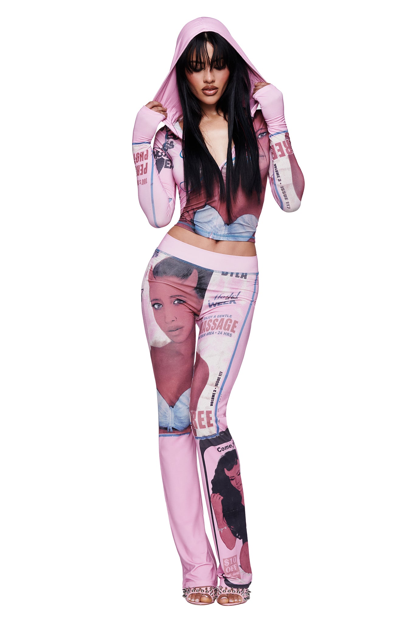 A woman wears the I.AM.GIA T-REX BLARE TRACKPANT in pink, styled with a matching hooded outfit adorned with illustrated faces, magazine-inspired graphics, and a striking T-REX pink print. With her hood up, she looks directly at the camera. The trackpants feature a wide elastic waistband for both comfort and style.