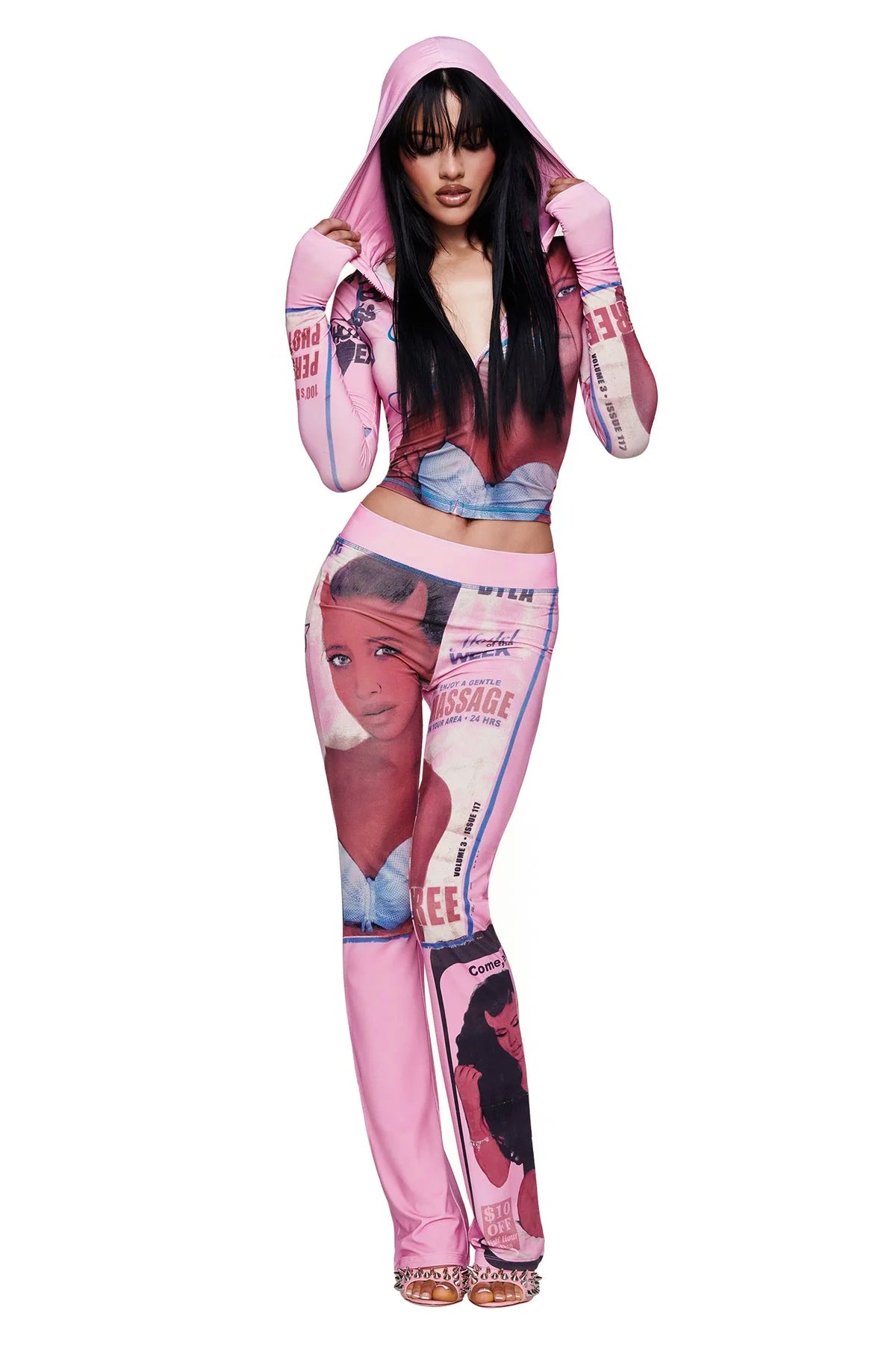 A woman stands with her hands holding the hood of her two-piece T-REX BLARE TRACKPANT set in pink by I.AM.GIA, featuring bold prints of women's faces and magazine covers. With long dark hair and bangs, she gazes forward, capturing the edgy style of this standout ensemble.