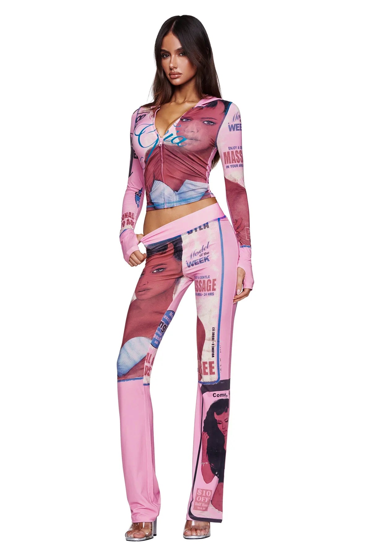 A woman stands against a white background wearing the I.AM.GIA T-REX BLARE HOODIE in pink, which features magazine-style graphics and faces. She has long brown hair, clear heels, and poses with one hand on her hip.