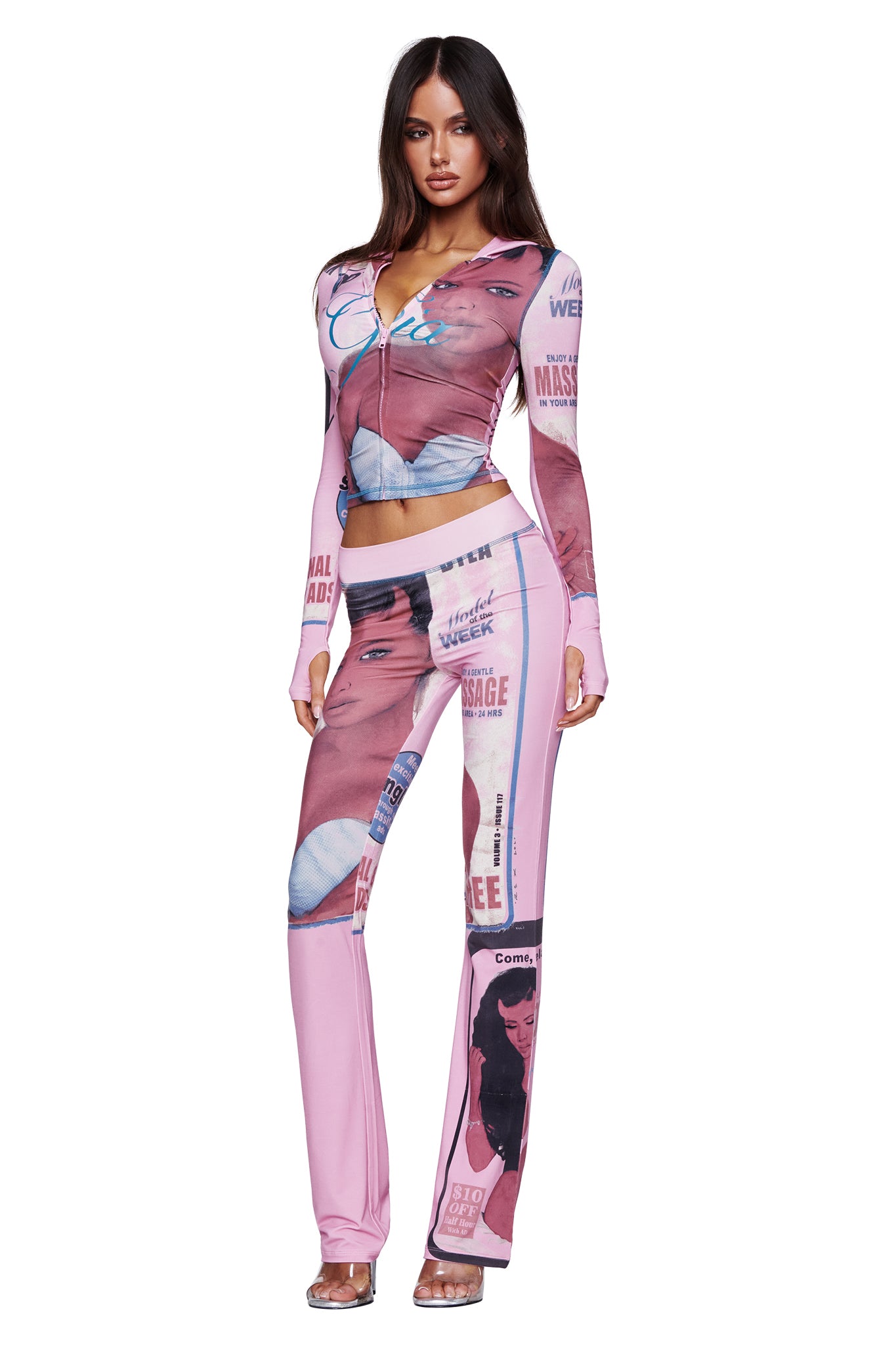A woman is wearing the I.AM.GIA T-REX BLARE TRACKPANT in pink, styled with a matching graphic zip-up crop top featuring T-REX pink print details, illustrated faces, and bold text. The wide elastic waistband of the flared pants ensures comfort, while she completes the look with clear high-heeled sandals.