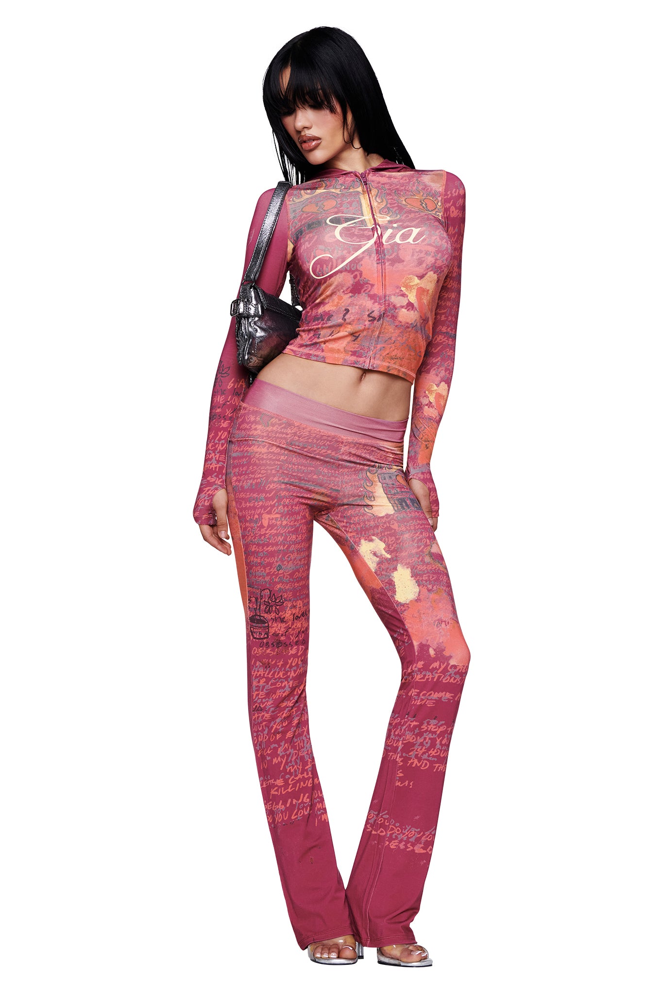 A woman poses in a pink and orange fitted ensemble with script text, made up of a long-sleeve crop top and matching pants—her striking look echoing the vibrant energy of I.AM.GIA’s T-REX BLARE HOODIE in red. She completes her outfit with a small black shoulder bag and heeled sandals.