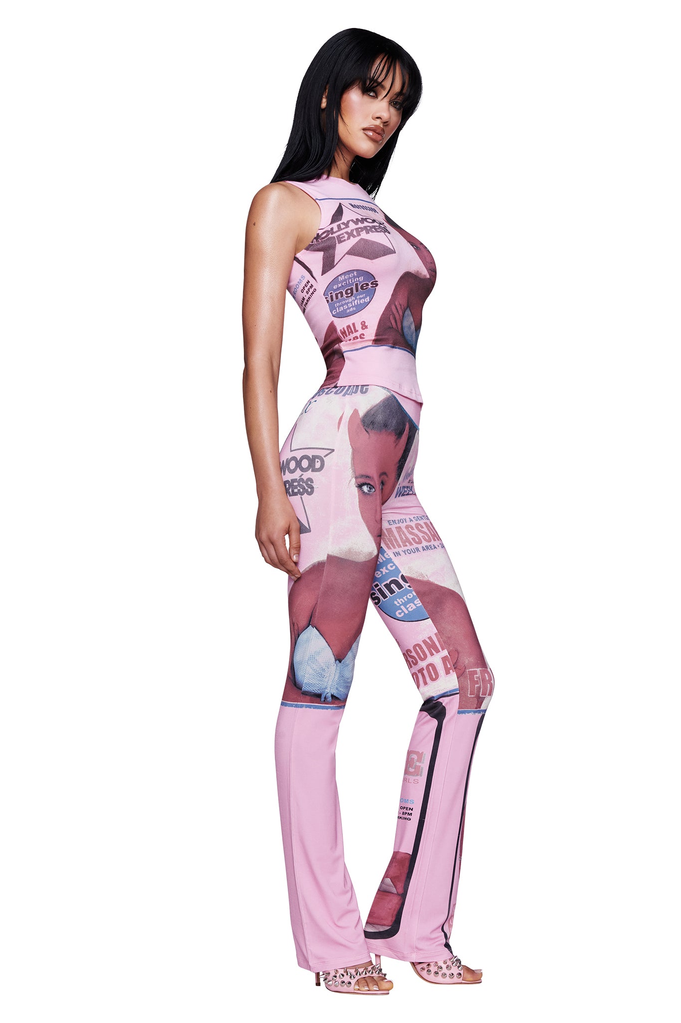 A woman stands sideways wearing the I.AM.GIA T-REX BLARE TRACKPANT in pink, featuring various vintage-inspired graphic prints—including a bold T-REX motif—paired with open-toe heels and long dark hair.