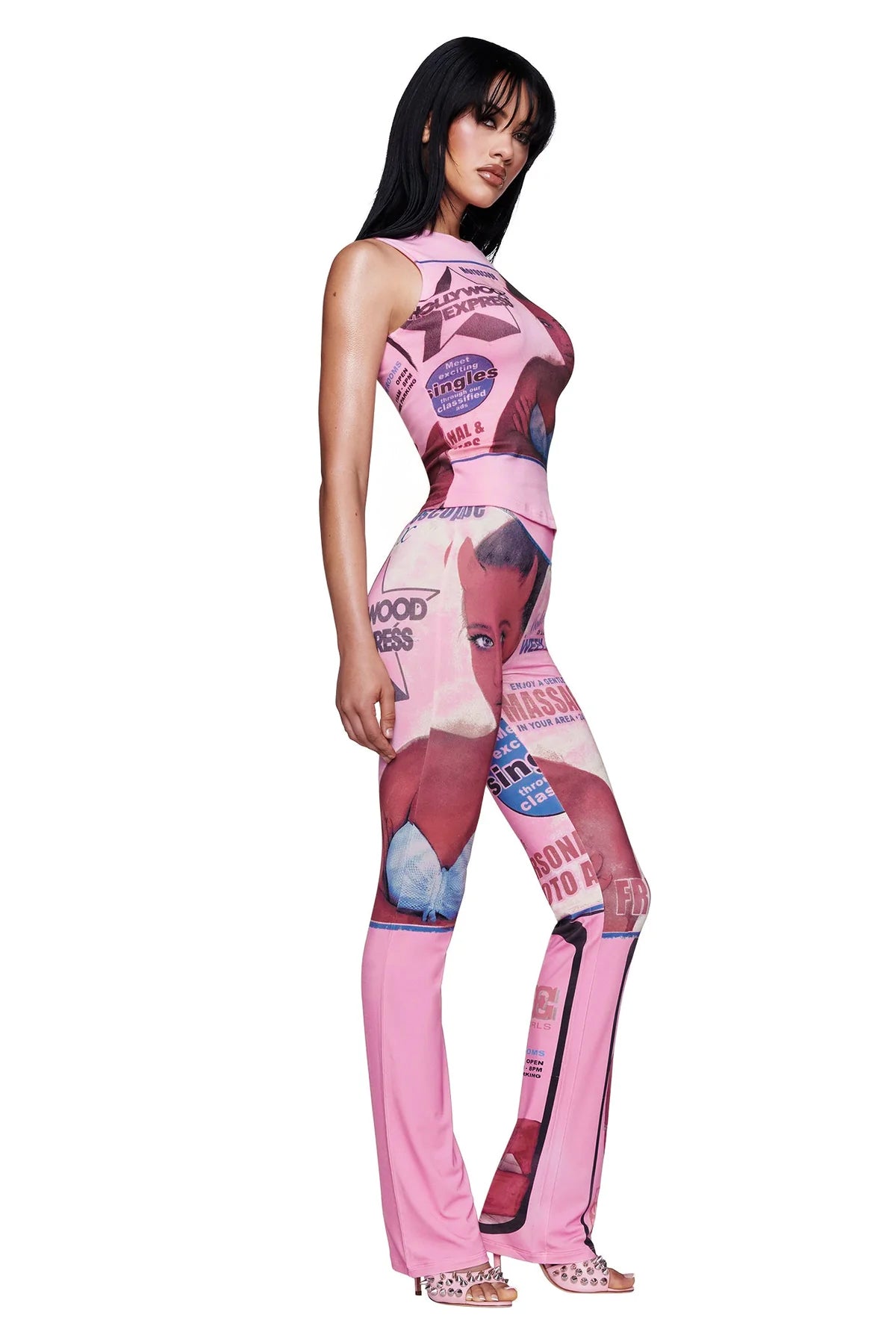A woman stands confidently in the T-REX BLARE TRACKPANT in pink by I.AM.GIA, featuring graphic images and text inspired by the signature T-REX print. She pairs the statement track pants with strappy heels, her long dark hair cascading as she poses against a white background, looking off to the side.