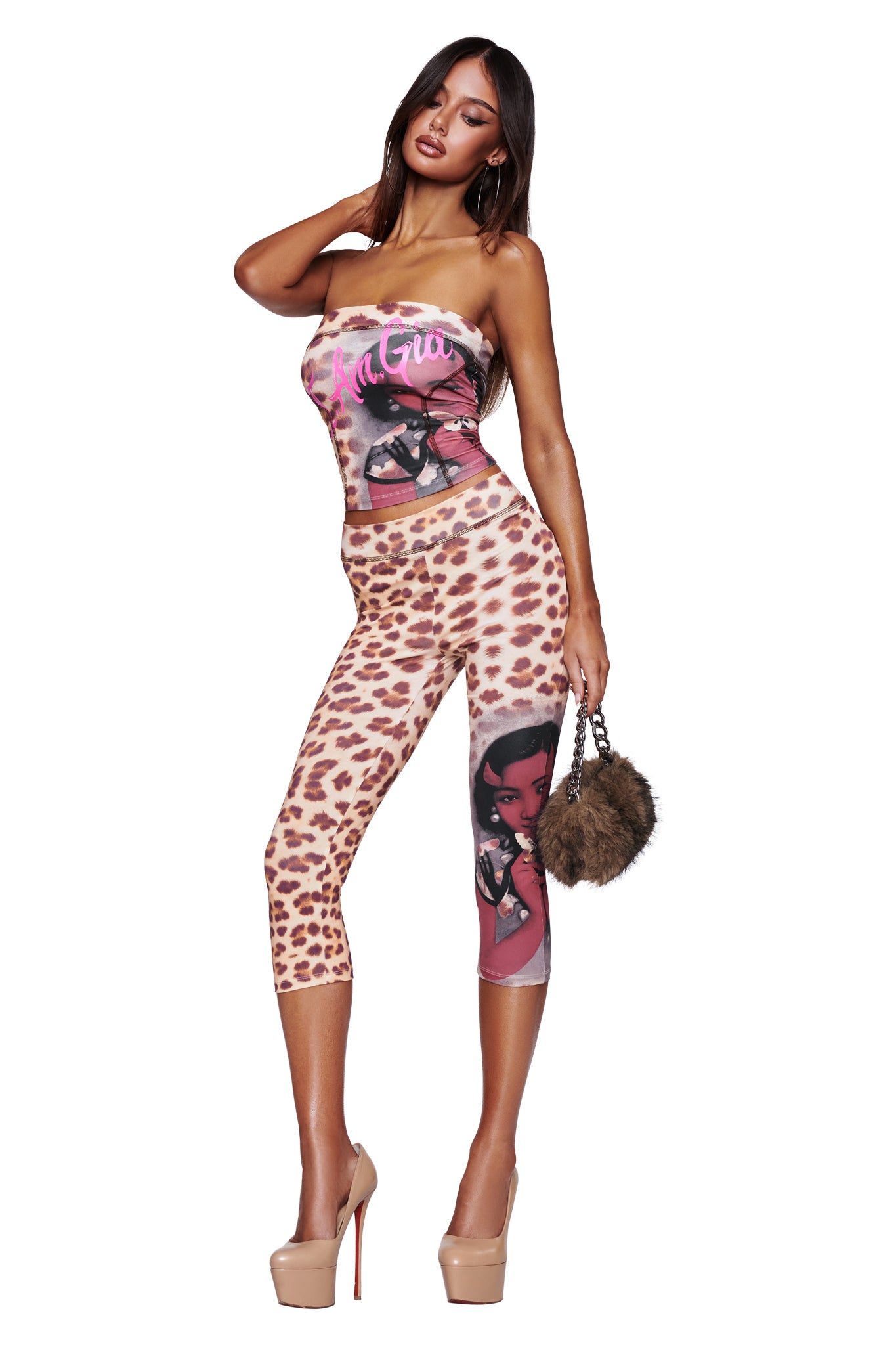 Wearing the I.AM.GIA T-REX BLARE BOOB TUBE - WILD CARD and matching leggings featuring a bold cheetah print with cartoon graphics, a woman poses against a white background. She accessorizes her look with a furry handbag and nude high heels.