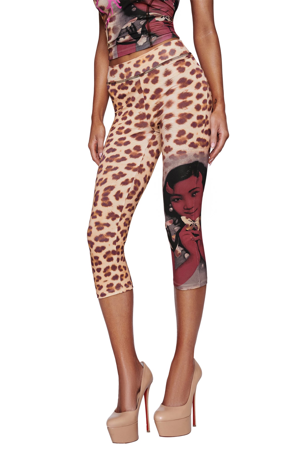 A model wears the I.AM.GIA T-REX BLARE CAPRI - WILD CARD, a cheetah print, bodycon-fit capri pant featuring a graphic of a woman's face on one leg, styled with nude high heels. The upper body and face are not visible against a white background.