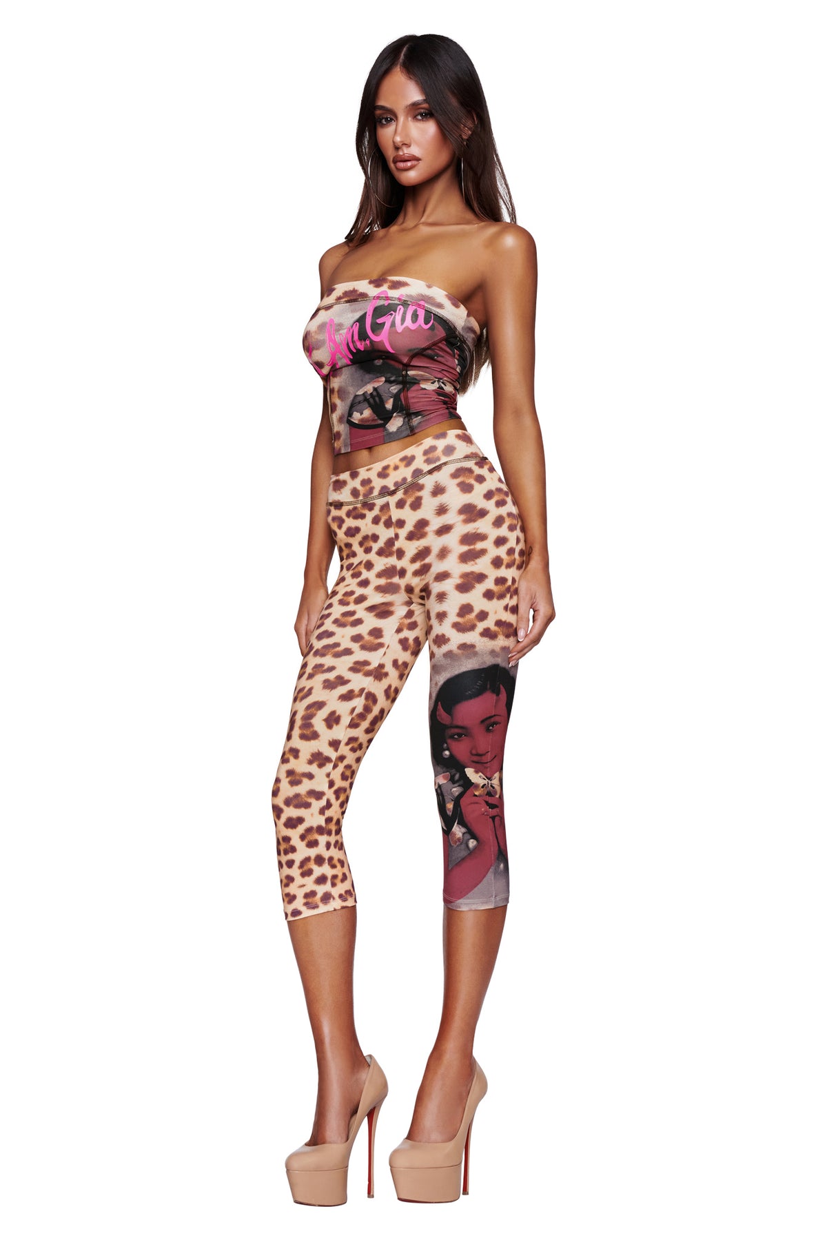 A woman wearing the I.AM.GIA T-REX BLARE CAPRI - WILD CARD cheetah print bodycon leggings featuring a bold cartoon graphic on one leg, paired with a coordinating leopard print strapless top and nude high heels, stands against a white background.
