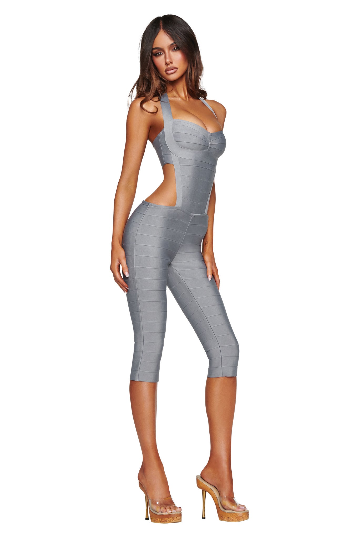 A woman poses in the I.AM.GIA EVA BANDAGE CAPRI JUMPSUIT in grey—a fitted, sleeveless style featuring chic cut-out details—paired with clear high-heeled shoes against a white background.