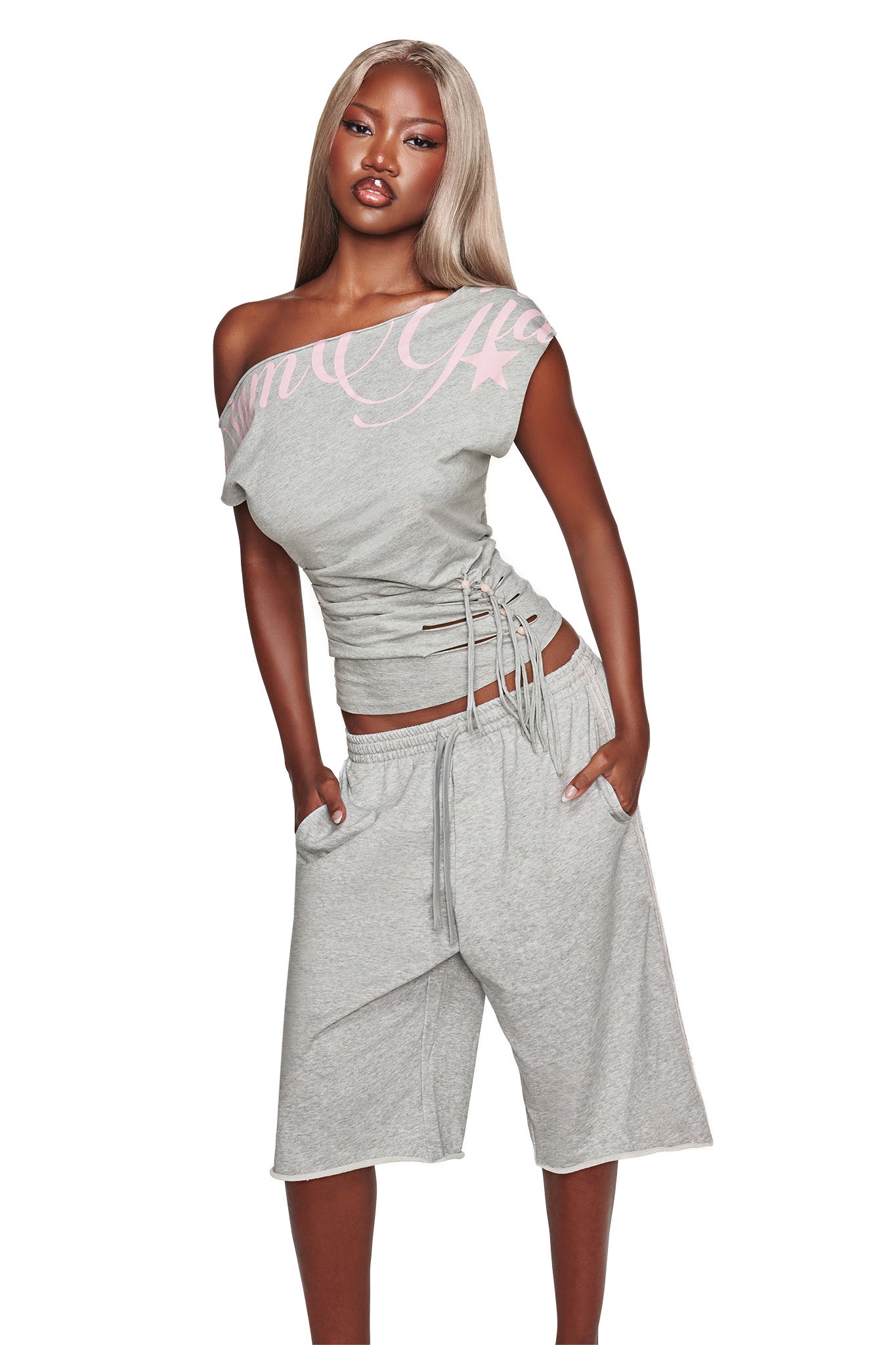 A woman with long blonde hair models the I.AM.GIA BEX TERRY SHORT in grey, featuring a relaxed fit and an elasticated waistband. She pairs the shorts with a gray off-shoulder top adorned with pink lettering and side ties, standing confidently with her hands in her pockets against a white background.