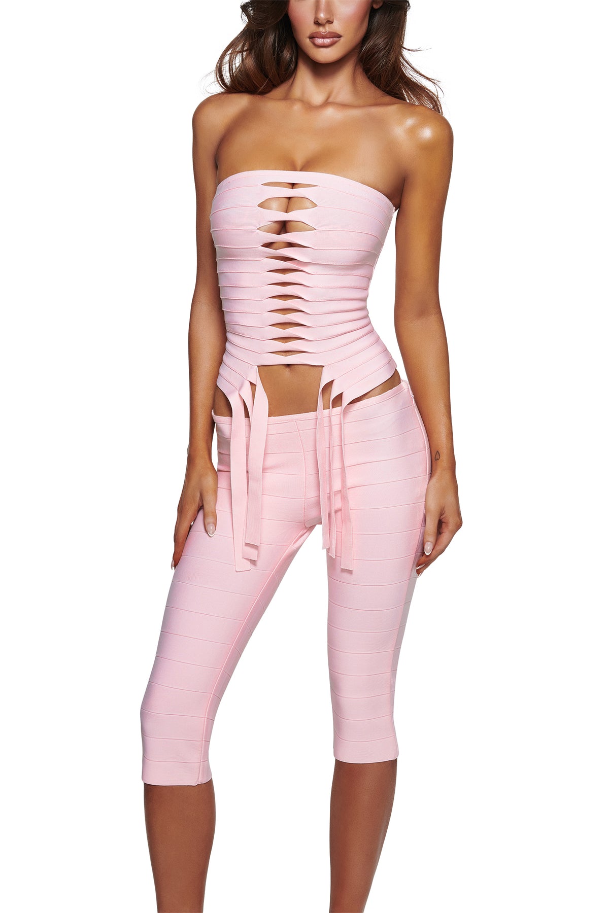 A woman stands against a white background wearing the MAEVE BANDAGE CAPRI - BABY PINK by I.AM.GIA, featuring a strapless, pale pink bandage top with center front cutouts and matching baby pink capri pants in a sleek bodycon fit.