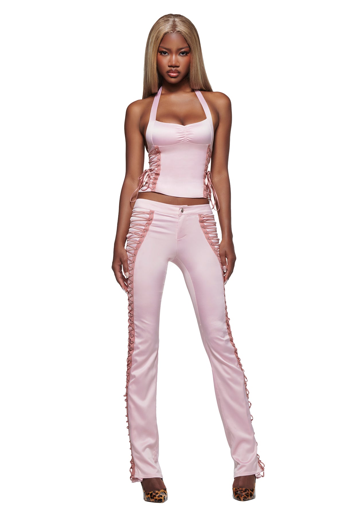 A woman stands facing forward in the I.AM.GIA HOLLI SATIN PANT 2.0 - PINK with matching light pink halter top, featuring side cutouts and lace-up detailing. She wears leopard print high heels and has long, straight blonde hair against a plain white background.