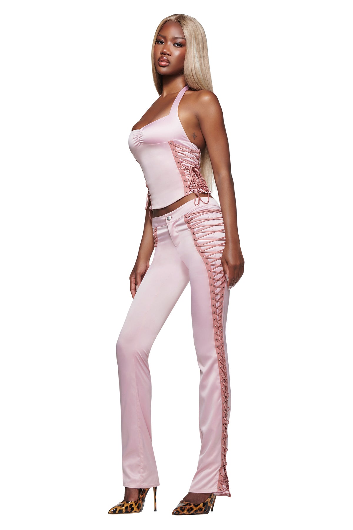 A woman with straight blonde hair is wearing the I.AM.GIA HOLLI SATIN CORSET in pink and matching pink pants, both featuring ribbon detail. She completes the look with leopard print high heels and is standing sideways against a white background.