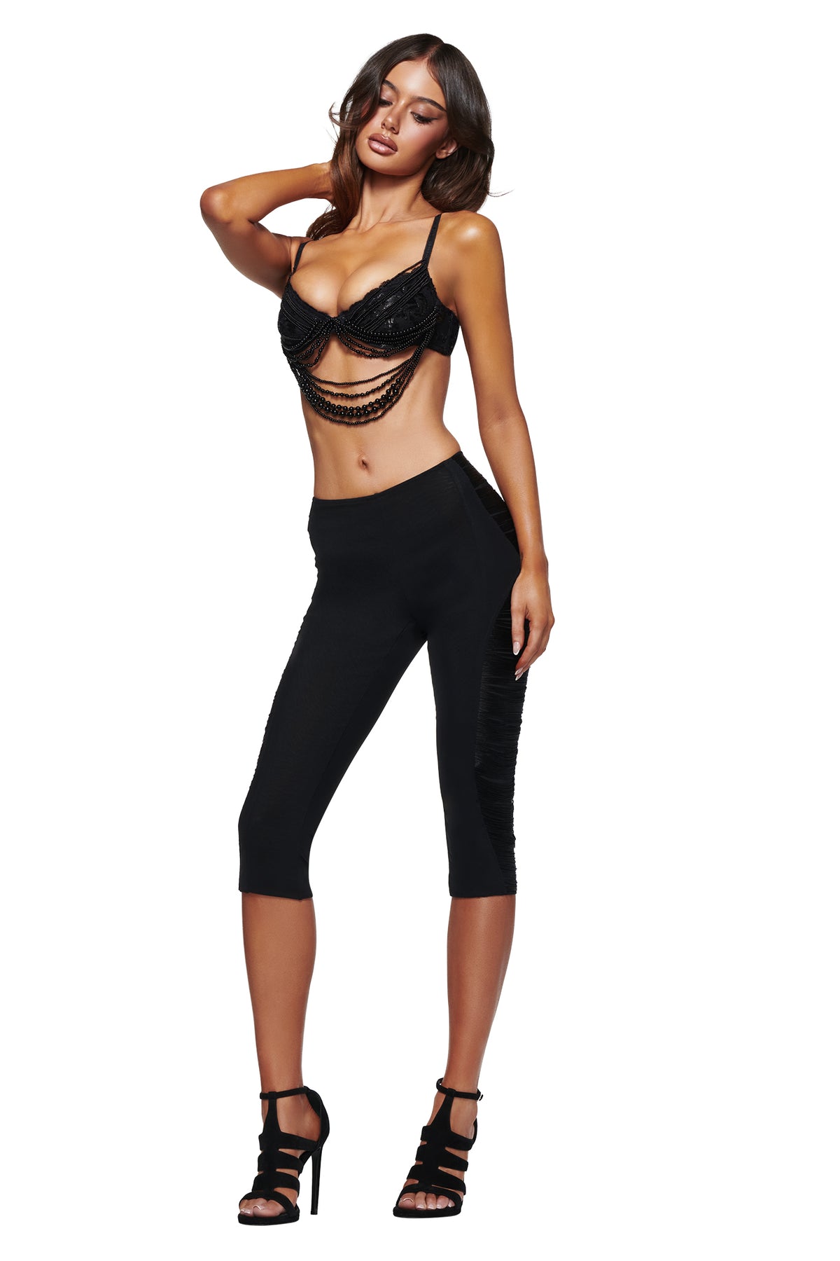 A woman wearing the I.AM.GIA VERON BRA in black, paired with black cropped pants and black high heels, poses with one hand behind her head against a white background.