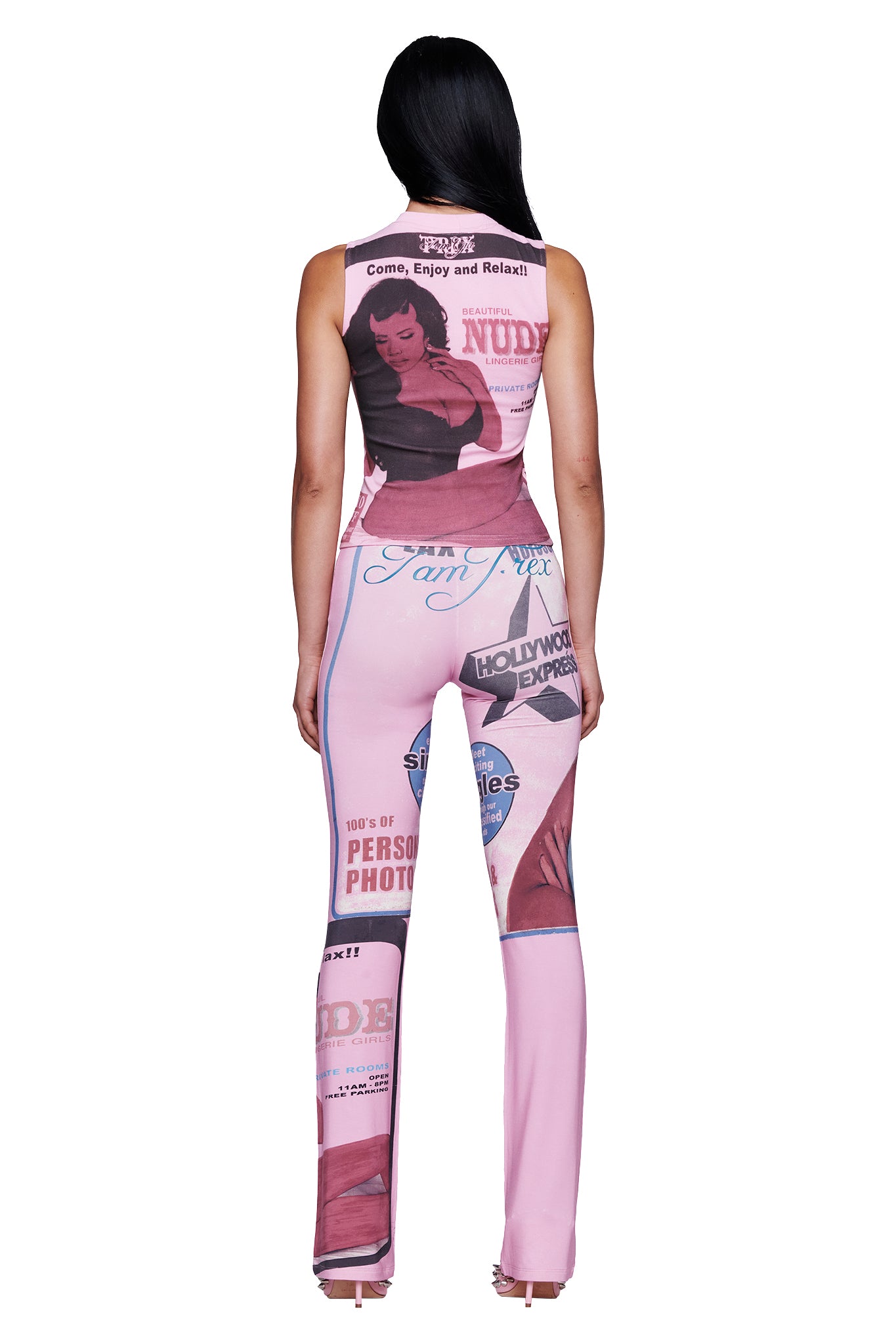 A woman stands facing away, wearing the I.AM.GIA T-REX COURTNEY TANK in pink—a sleeveless, form-fitting top reminiscent of bold muscle tank styles—printed with retro magazine covers, striking headlines, and a vintage pin-up illustration. She has long, straight black hair and is paired with open-toed heels.