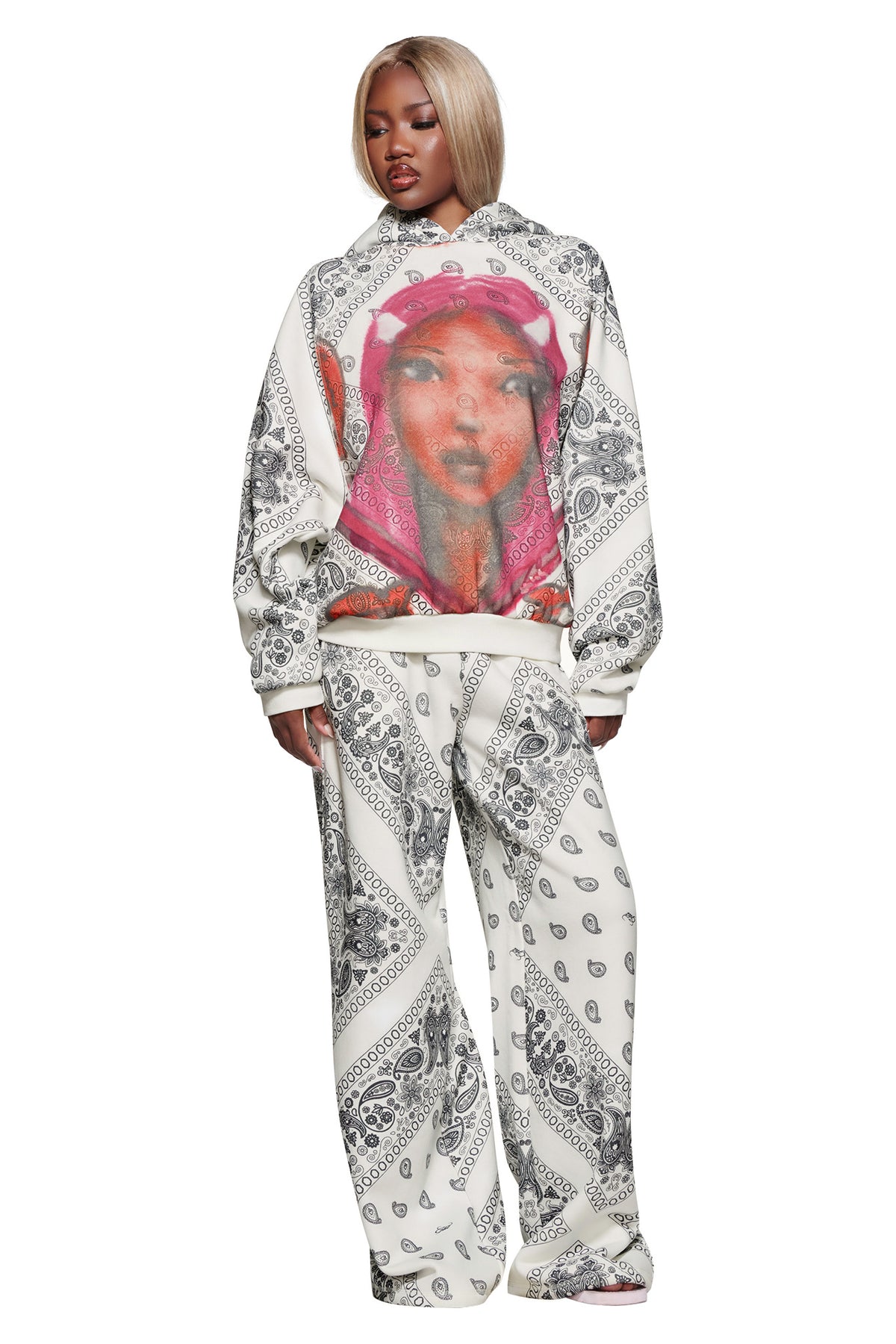 A person wears loose white pants and the I.AM.GIA T-REX PULLOVER HOODIE - PAISLEY FACE, with black bandana patterns and a large graphic of a woman’s face in a pink hood on the front.