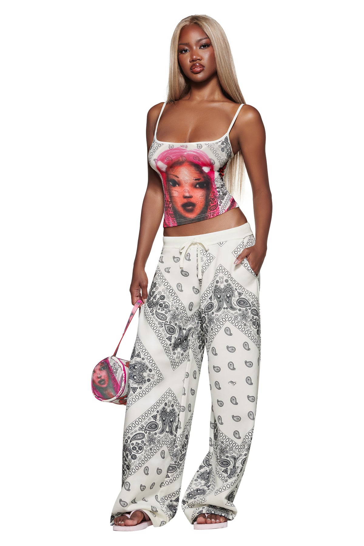Woman with long blonde hair wears a graphic tank top, I.AM.GIA T-REX TRACKPANT - PAISLEY, and holds a small pink purse. She stands confidently against a white background.