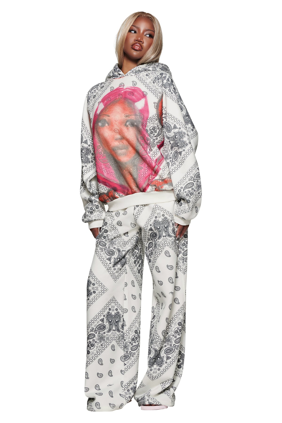 A woman wears the I.AM.GIA T-REX TRACKPANT - PAISLEY and a matching white and black paisley bandana-print hoodie with a bold graphic of a woman in a pink bandana.