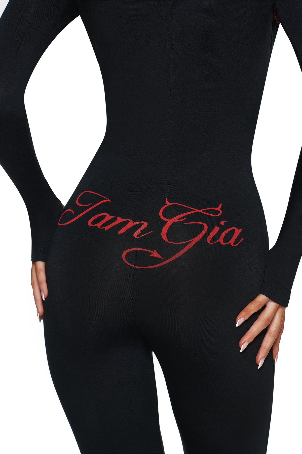 Person wears the BLARE HORN JUMPSUIT - BLACK by I.AM.GIA, featuring red script on the lower back, a playful devil horn hood, and white-painted nails resting on their hip.