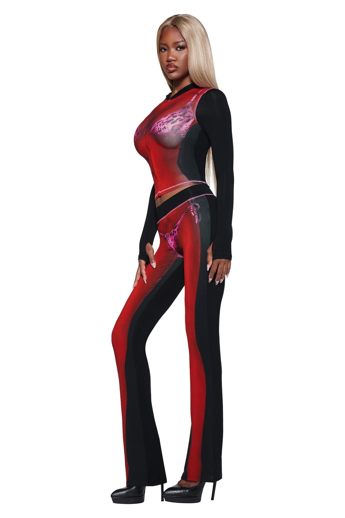 A woman with long blond hair models the I.AM.GIA T-REX SPRAY BODY BLARE HOODIE - BLACK, paired with matching pants and black heels, standing in profile against a white background.
