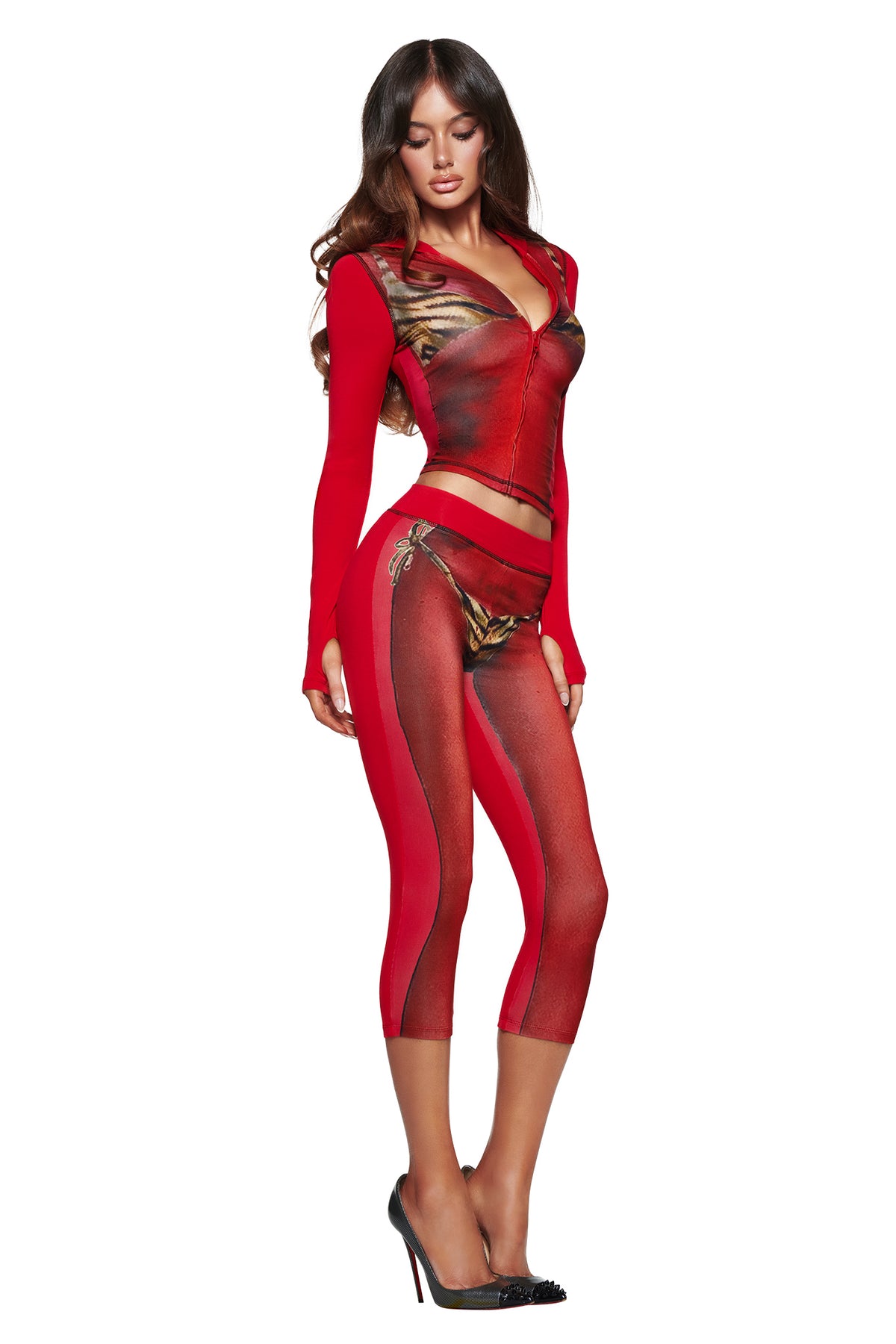 A woman in an I.AM.GIA T-REX SPRAY BODY BLARE HOODIE - RED, crop top, and leggings with an airbrushed bikini print pairs the look with black heels, posing confidently against a white background.