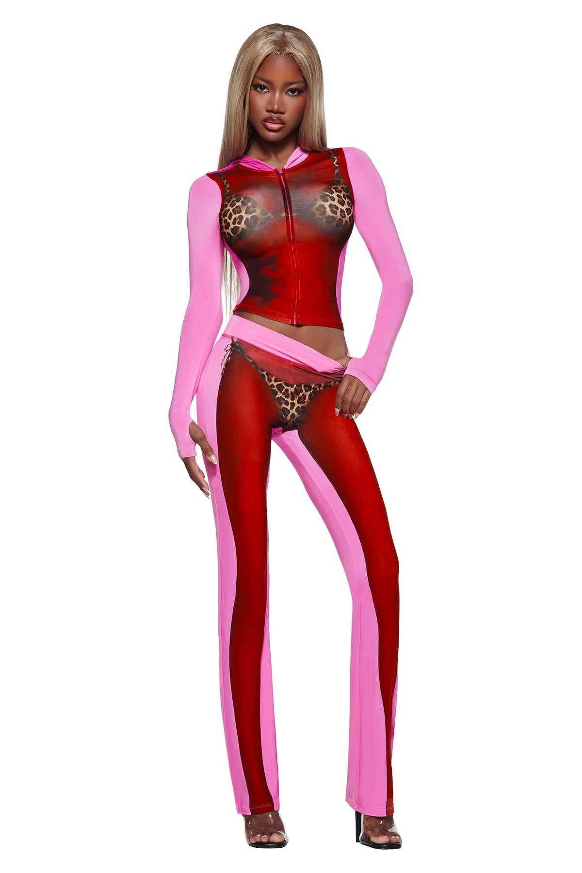 A woman with long blonde hair wears the T-REX SPRAY BODY BLARE TRACKPANT - PINK by I.AM.GIA, styled with a sheer red top and black heels—perfect for bold airport fashion against a white background.
