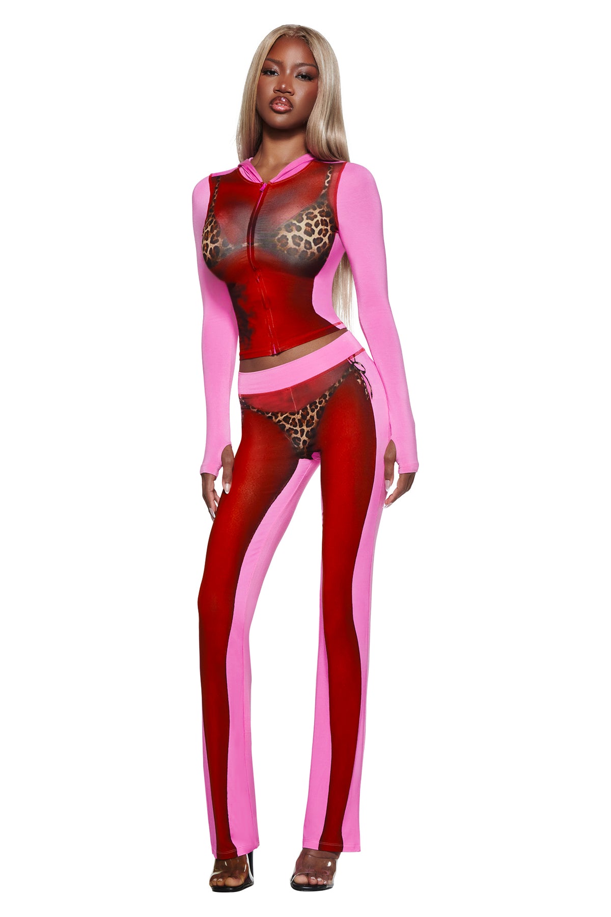 A woman poses in the I.AM.GIA T-REX SPRAY BODY BLARE TRACKPANT - PINK with sheer panels and leopard-print details, styled with long sleeves, a bikini set, and heels against a white background.
