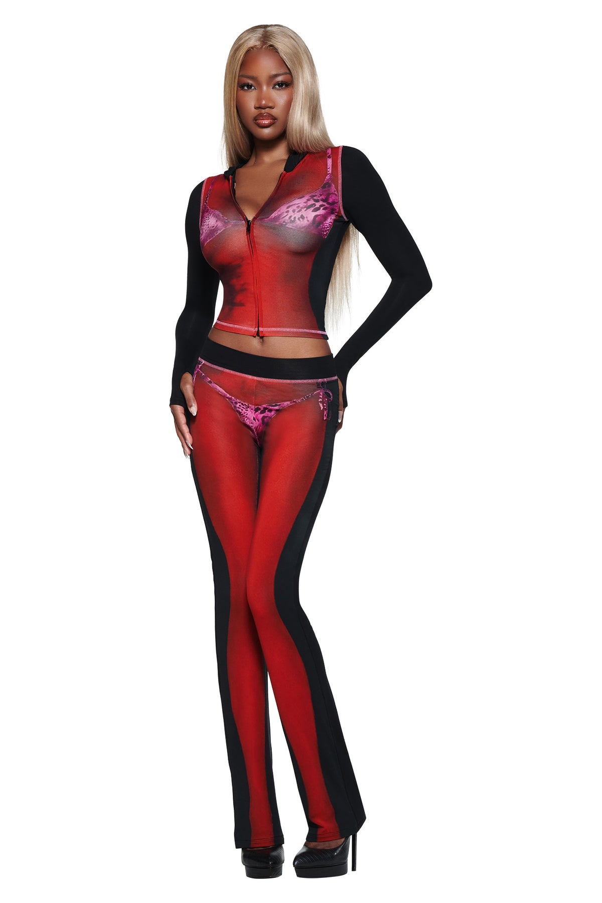 A woman with long blonde hair models the I.AM.GIA T-REX SPRAY BODY BLARE TRACKPANT - BLACK with a red and black sheer zip-front top, revealing red underwear, and poses confidently in black heels against a white background.