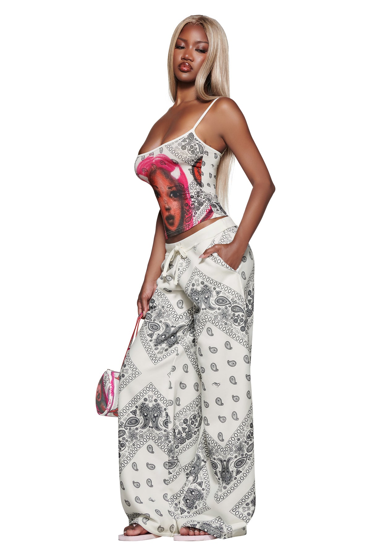 A woman with long blonde hair wears the I.AM.GIA T-REX TANK - PAISLEY FACE, loose white paisley pants, sandals, and holds a matching bag, standing with one hand in her pocket against a white background.