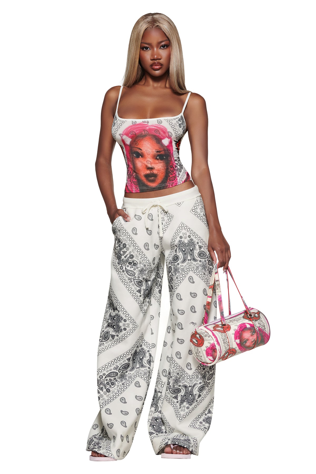 Woman with long blonde hair wears the I.AM.GIA T-REX TANK - PAISLEY FACE and matching pants, holding a coordinating bag, standing against a white background.
