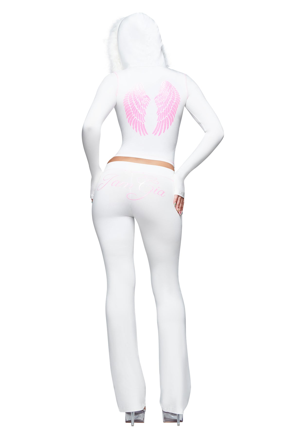 Dressed in the I.AM.GIA BLARE TRACKPANT - WHITE : ANGEL, featuring pink angel wings printed on the back and script detailing, this individual stands on clear platform heels with one hand on their hip. The low-rise flared trackpants and signature design elements embody the iconic I.AM.GIA ANGEL aesthetic.