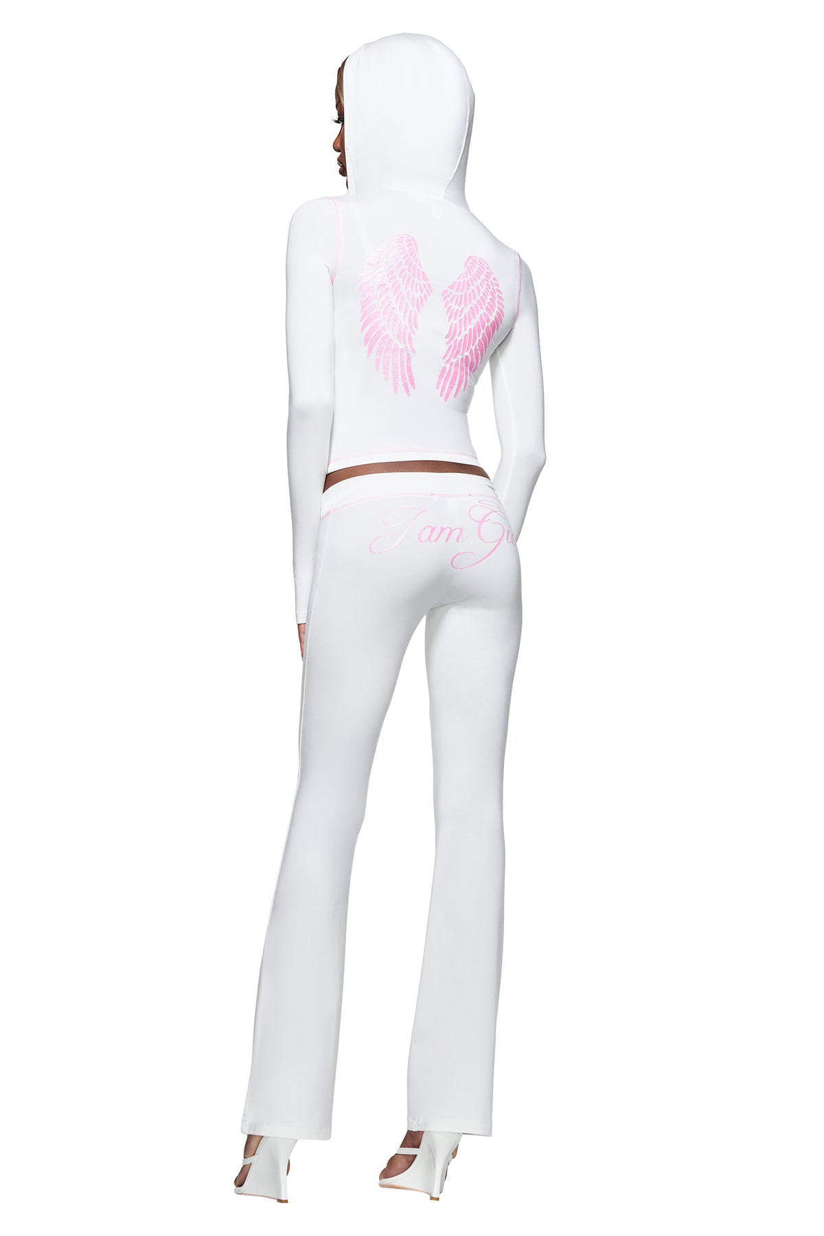 A person wearing the I.AM.GIA BLARE FUR TRIM HOODIE in white, featuring pink angel wings on the back, paired with pants that have “I am G” written on them and white high-heeled shoes, stands with their back to the camera.