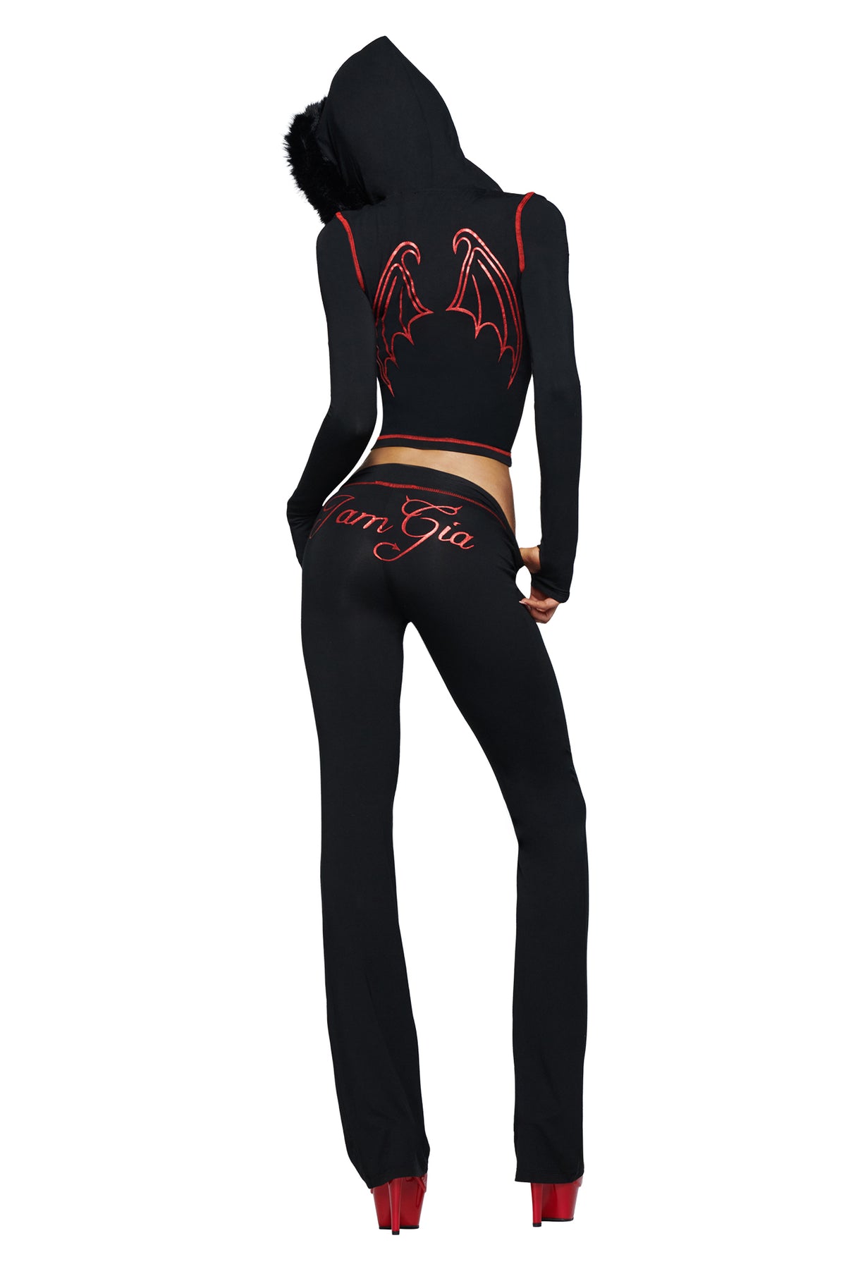 Model stands facing away from the camera, dressed in a black hooded outfit featuring red bat wings and the devil GIA logo on the back. She wears I.AM.GIA’s BLARE TRACKPANT - JET BLACK : DEVIL, emblazoned with “I AM GIA” lettering down the leg, paired with bold red platform heels.