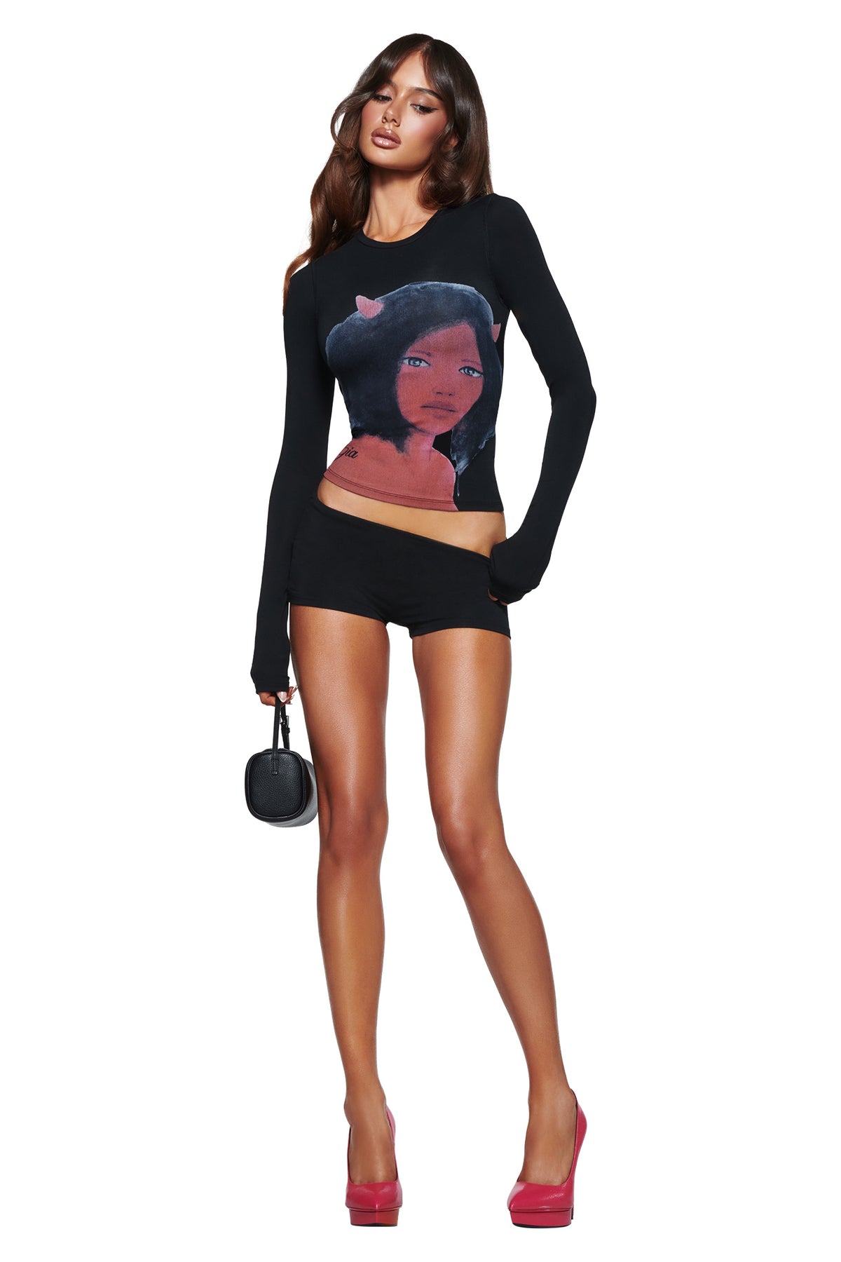 A woman in the T-REX BLARE 2.0 LONG SLEEVE TEE - BLACK by I.AM.GIA, black shorts, and pink heels poses with a small black bag against a white background.