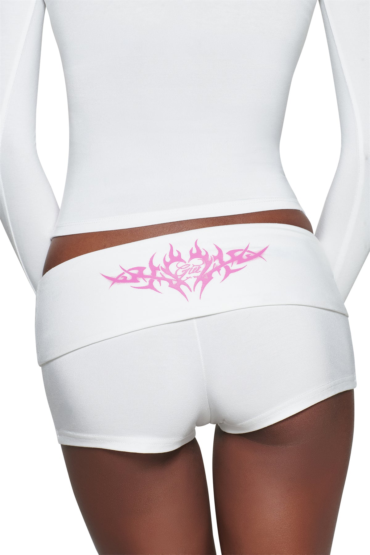 A person wearing white long-sleeve top and T-REX BLARE 2.0 SHORT - WHITE by I.AM.GIA, seen from the back. The mini shorts feature a pink tribal-inspired placement print across the foldover waistband.