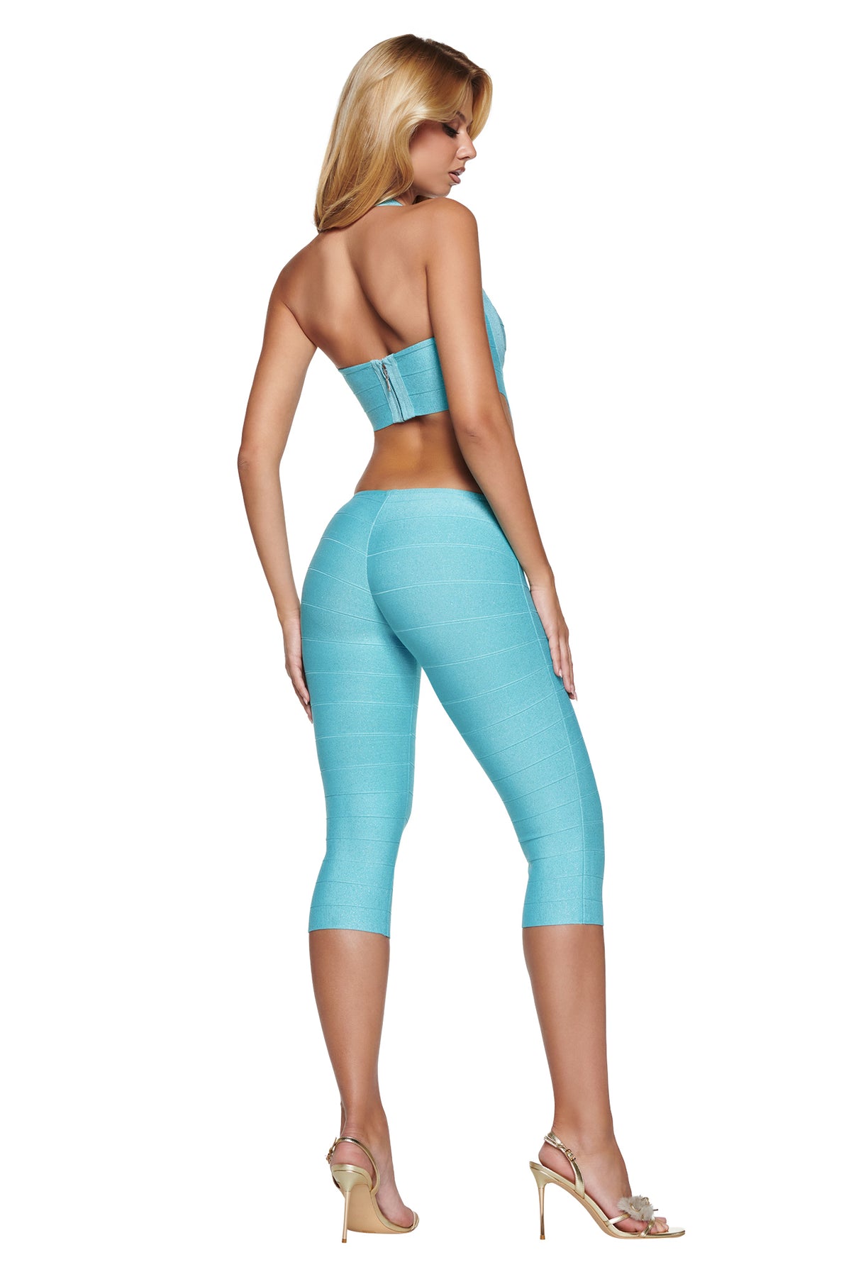 A person wearing an EVA SHIMMER BANDAGE CAPRI JUMPSUIT - METALLIC BLUE featuring a strapless crop top and matching bandage-style capri leggings paired with gold high-heeled sandals.